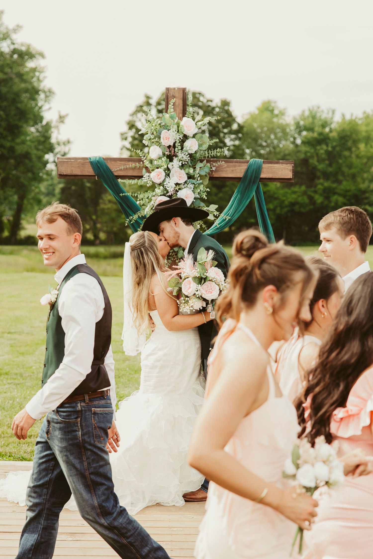 Bride and groom share first kiss at outdoor rustic cross altar surrounded by wedding party in jeans and pink bridesmaid dresses.