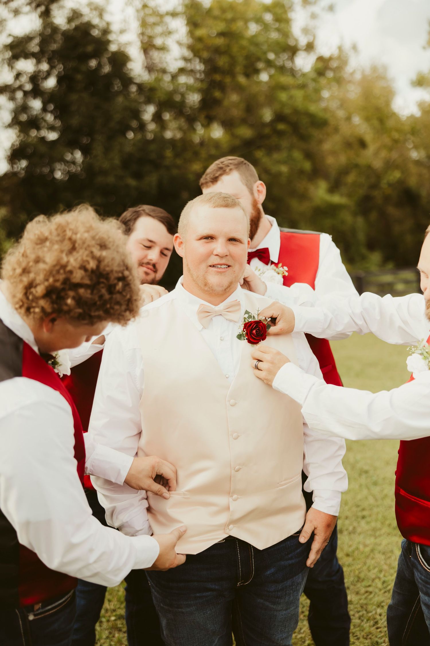 Groomsmen in red vests help groom with boutonniere outdoors before wedding ceremony.