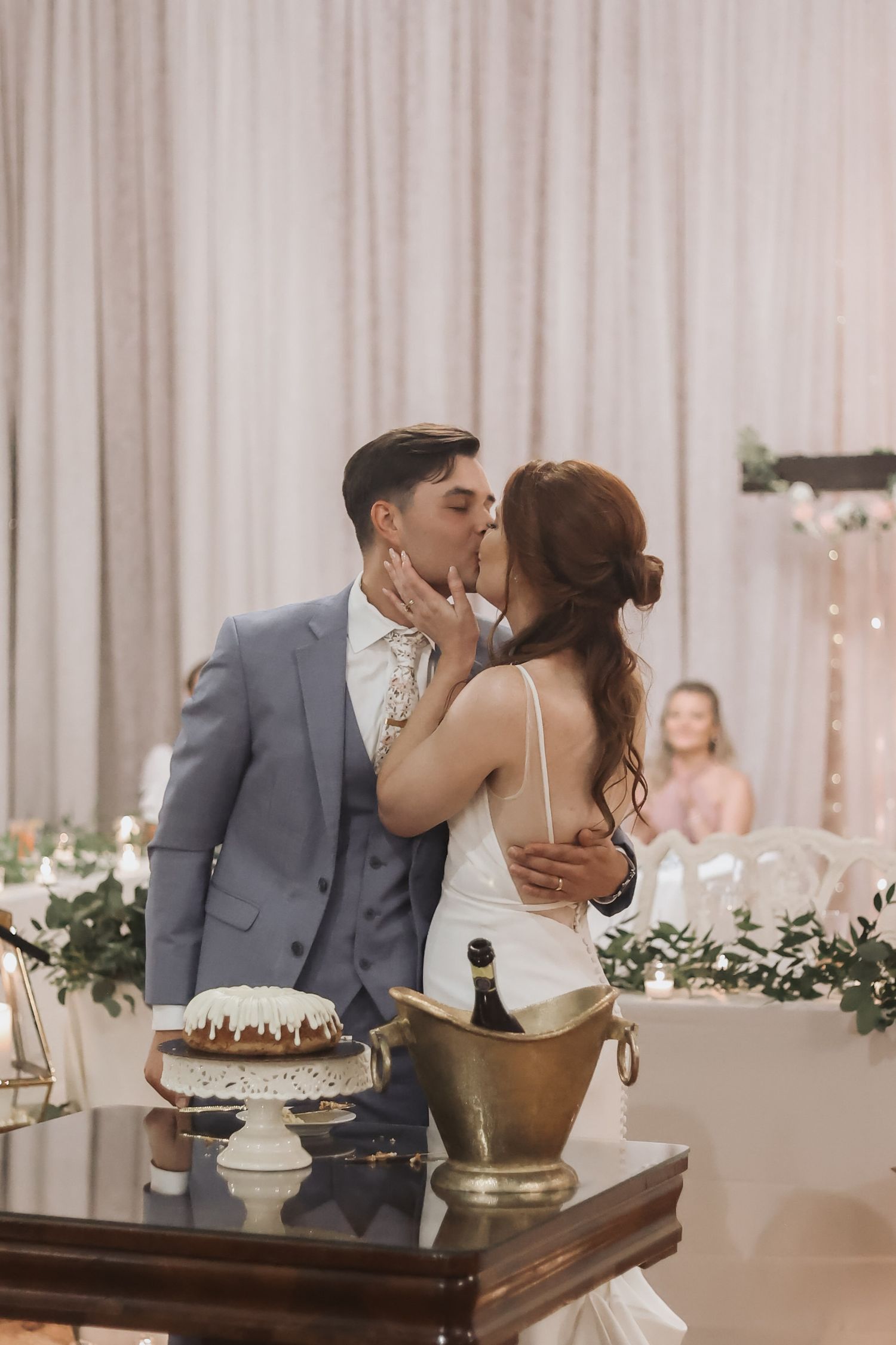 Newlyweds share a romantic kiss beside their wedding cake during an indoor reception with floral decorations.