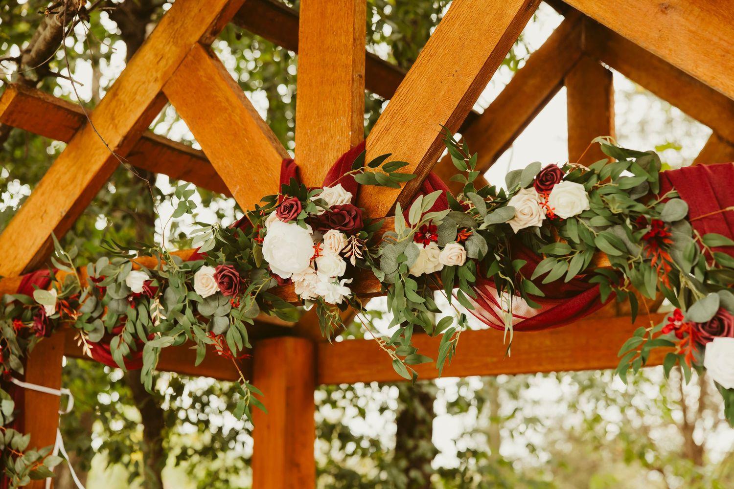 Rustic wooden wedding arch adorned with lush greenery, white roses, and burgundy blooms for an elegant outdoor ceremony.