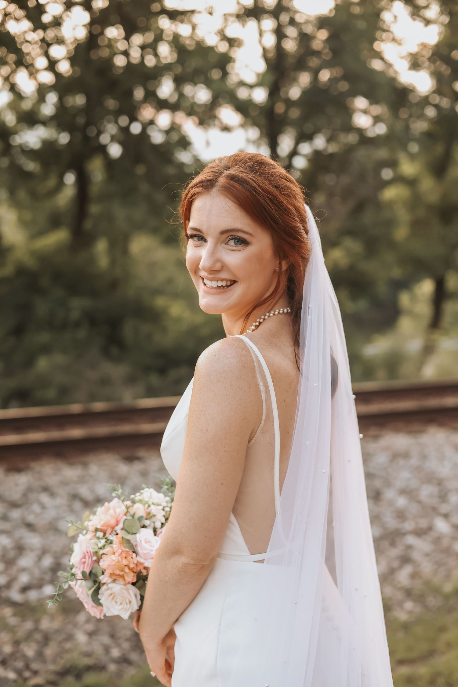 A bride in a white dress poses with her bouquet at sunset near railroad tracks.