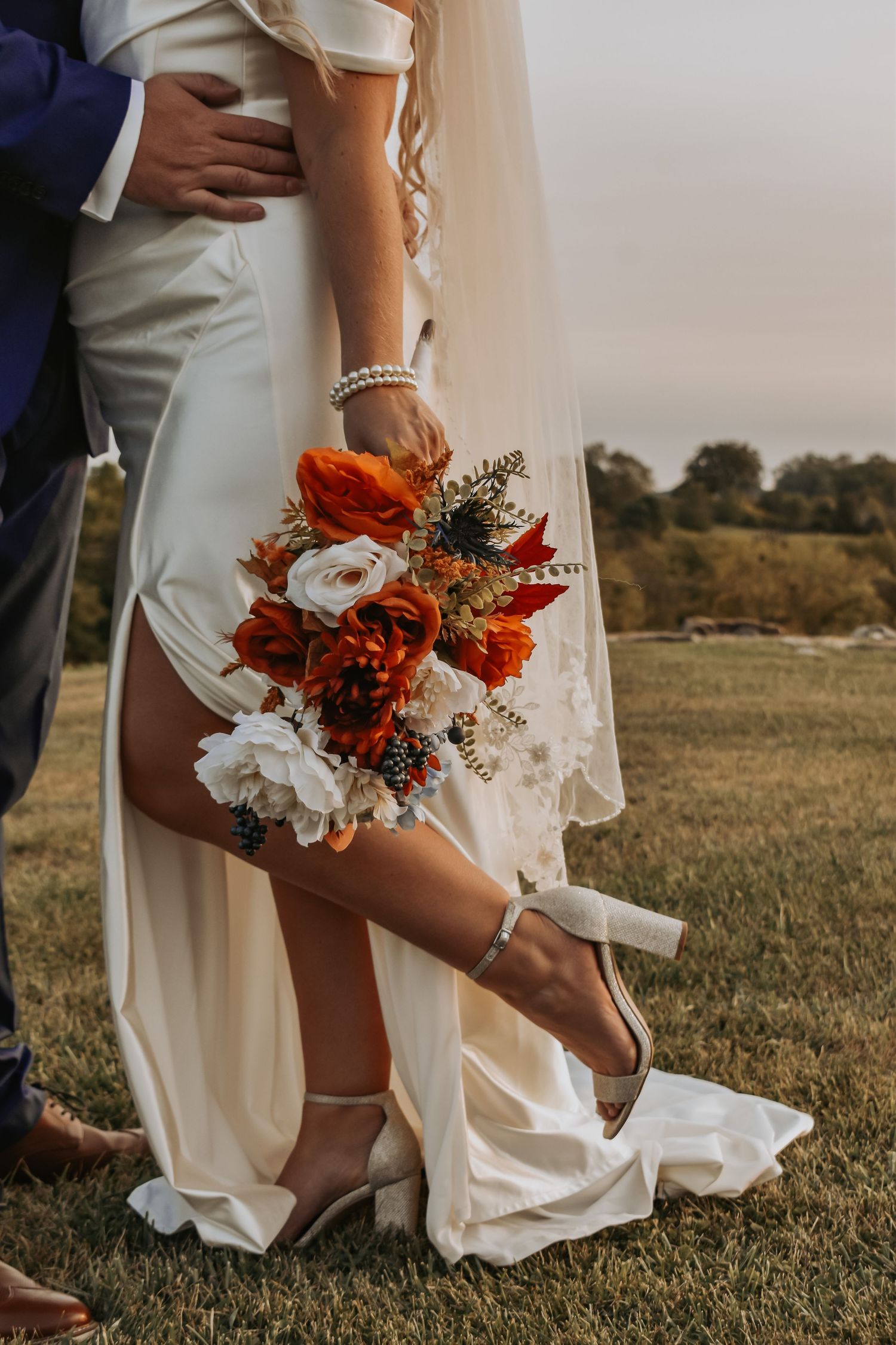 Close-up of bride's white dress and vibrant orange and white bouquet against a sunset landscape.