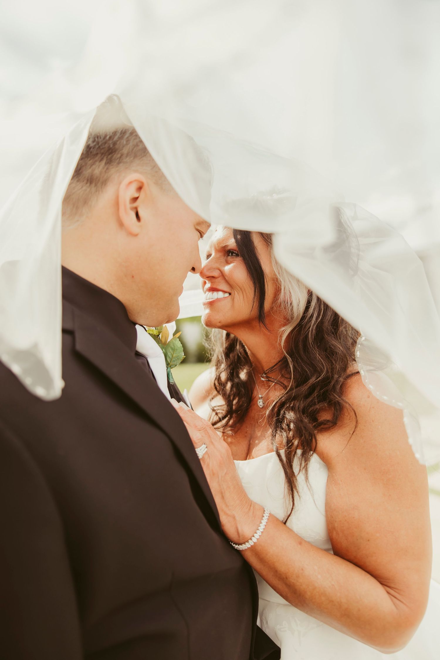 Romantic bride and groom nearly kissing under flowing white veil, bride smiling in strapless wedding gown.