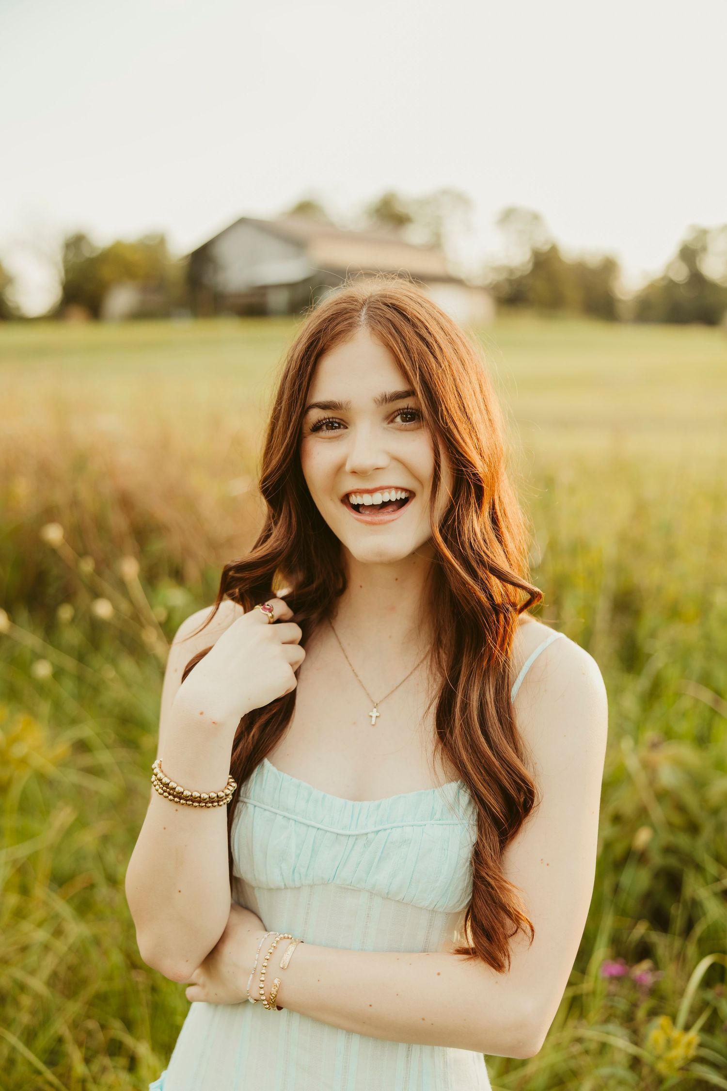 Young woman with long auburn hair smiling in a field, wearing a mint dress and gold jewelry at golden hour.