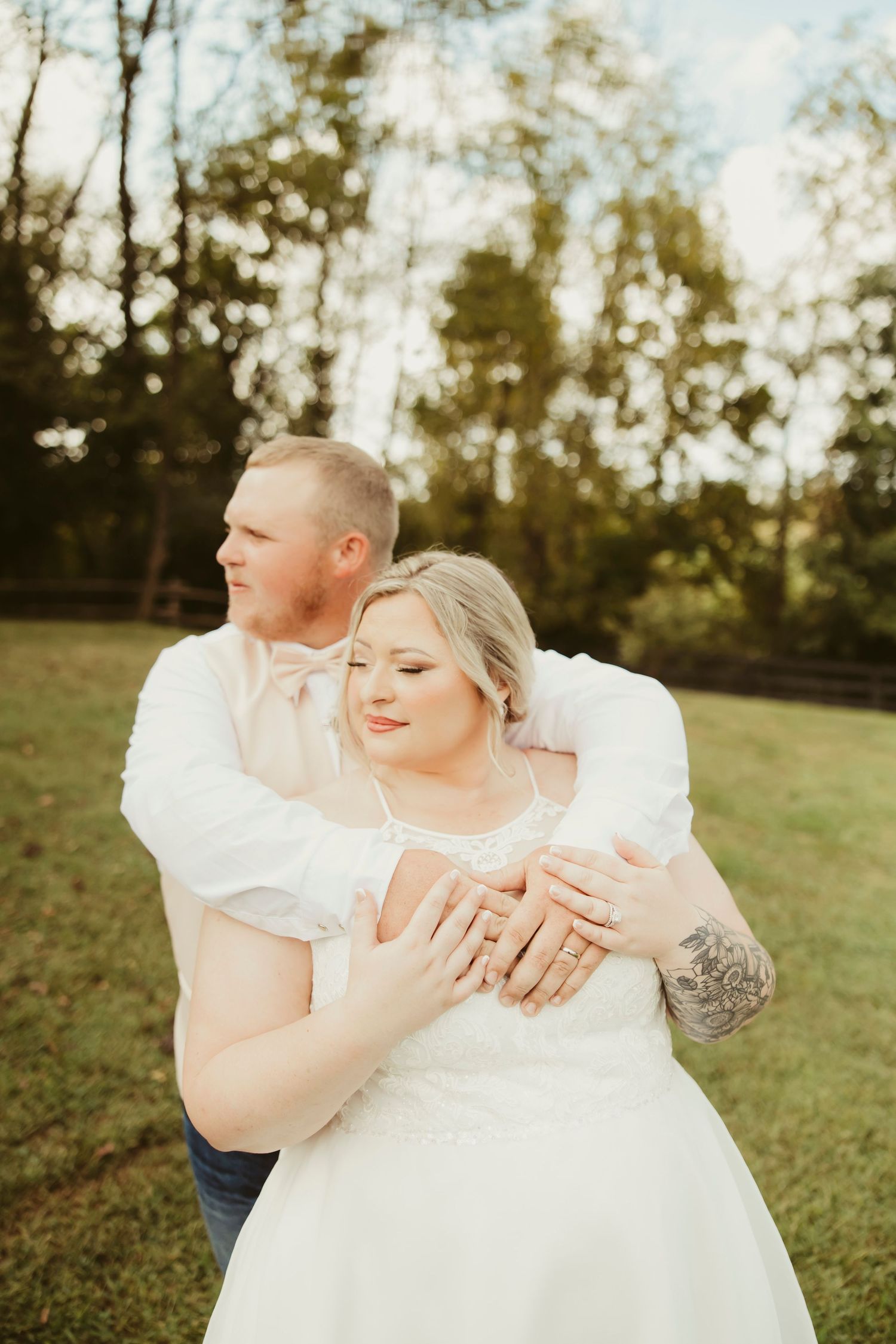 Bride and groom embrace outdoors in a green field, she wears a white lace dress as he holds her close.