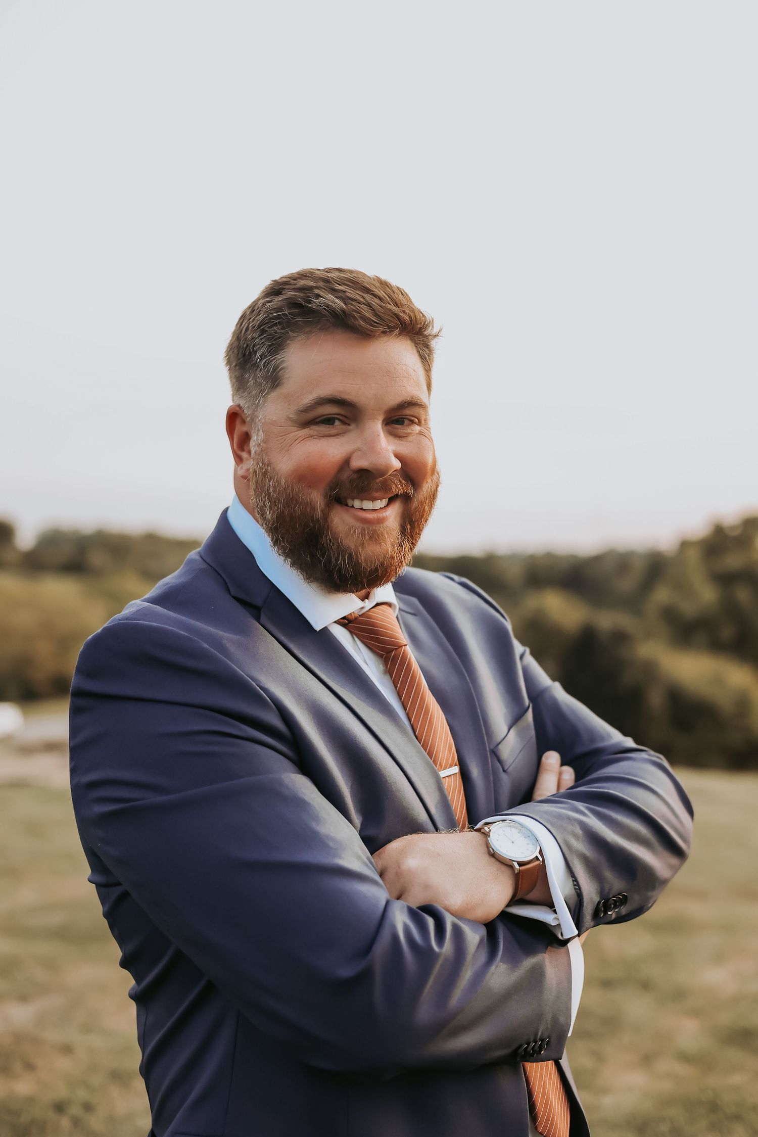 Groom in navy suit smiles confidently with arms crossed outdoors.