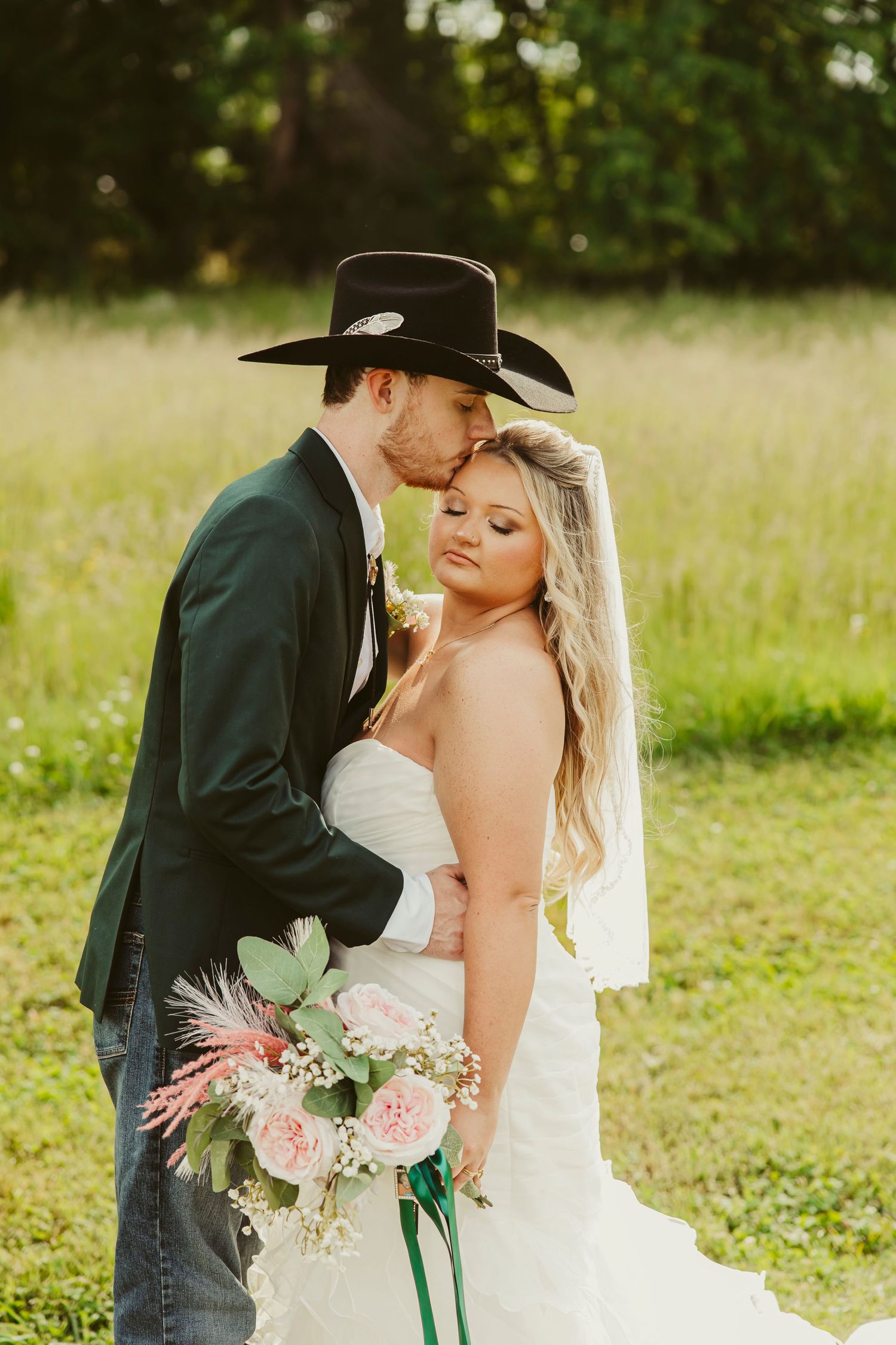 Groom in black cowboy hat kisses bride holding pink bouquet in a sunlit field during rustic country wedding.