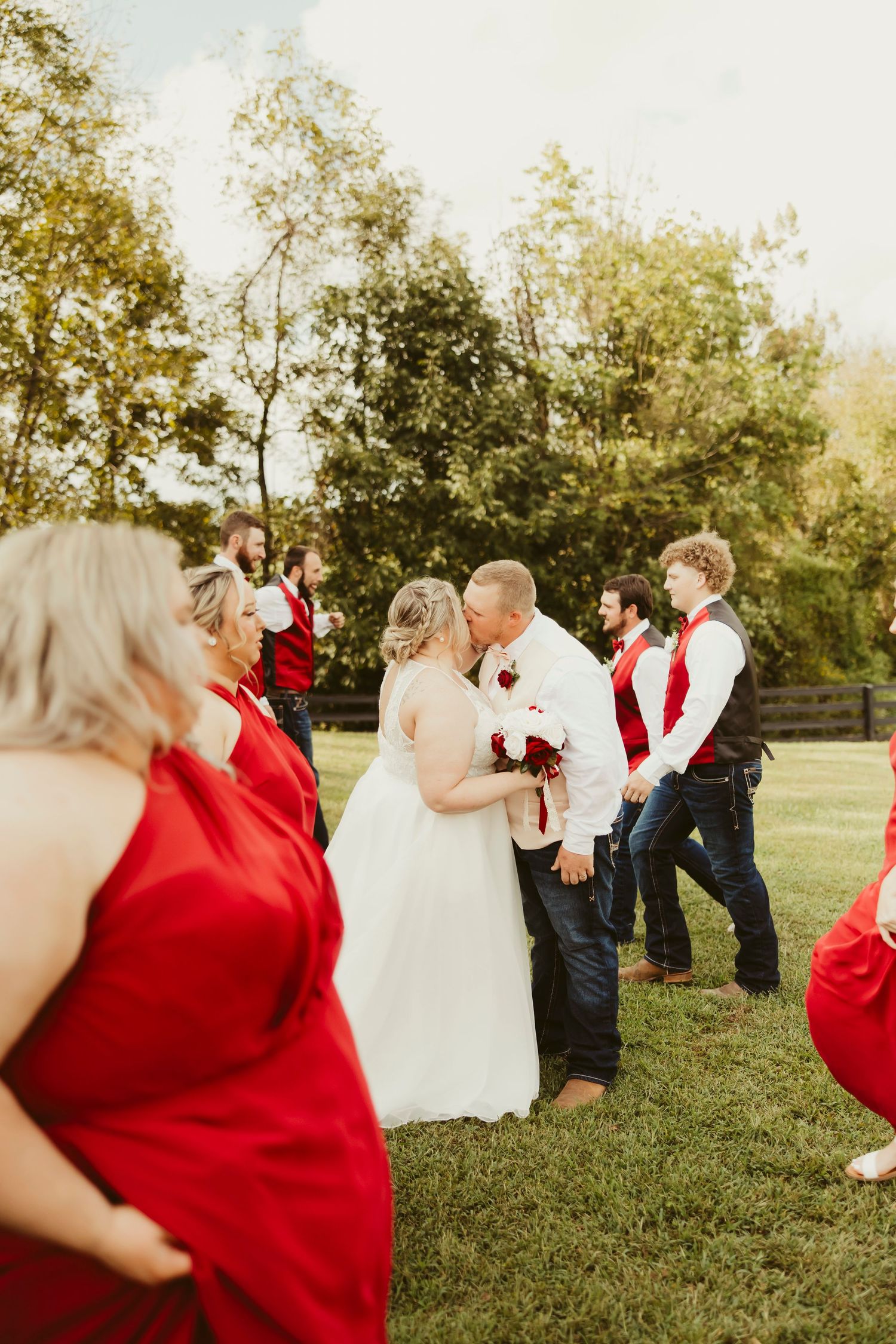 Bride and groom share a kiss outdoors surrounded by wedding party in red dresses and blue jeans on a sunny day.