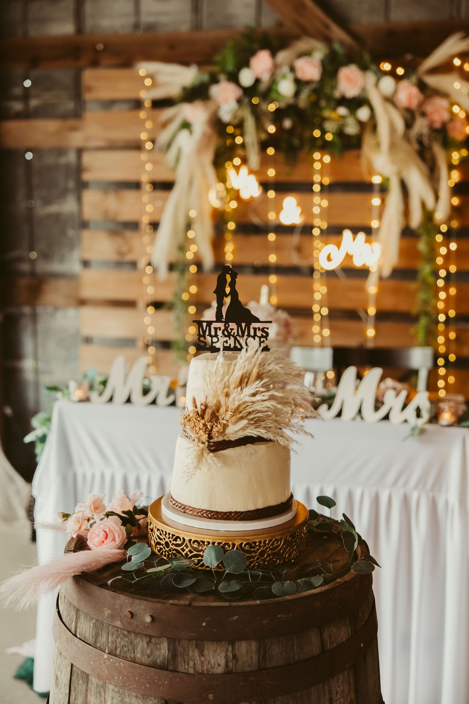 Rustic wedding cake with pampas grass topper on a barrel, framed by a pallet backdrop with fairy lights and floral decor.