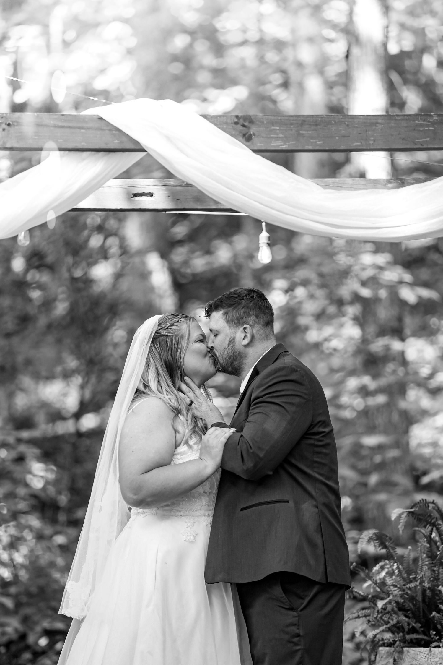 A romantic wedding kiss under a draped white fabric arch in a wooded outdoor setting, captured in black and white.