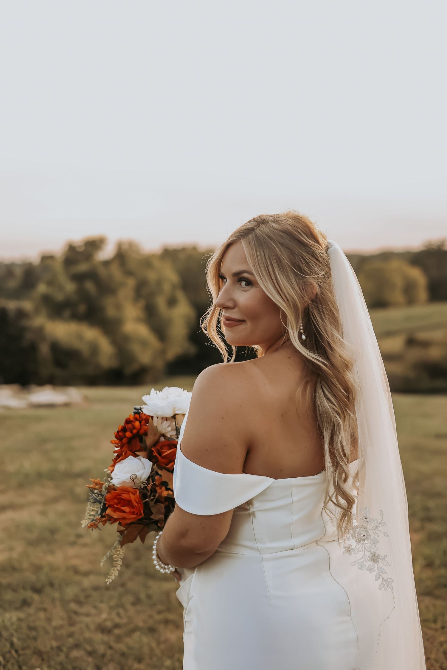 Bride with flowing veil holds orange and white bouquet in golden hour light.
