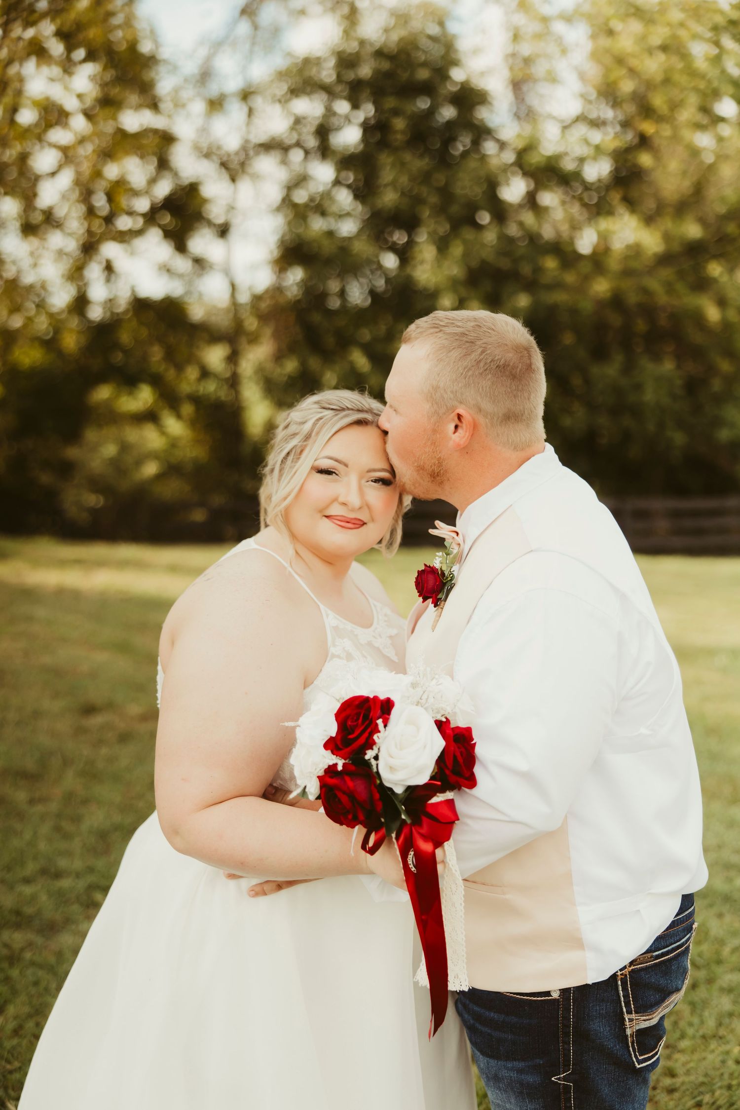 Bride holding red and white bouquet smiles as groom kisses her cheek outdoors on wedding day.