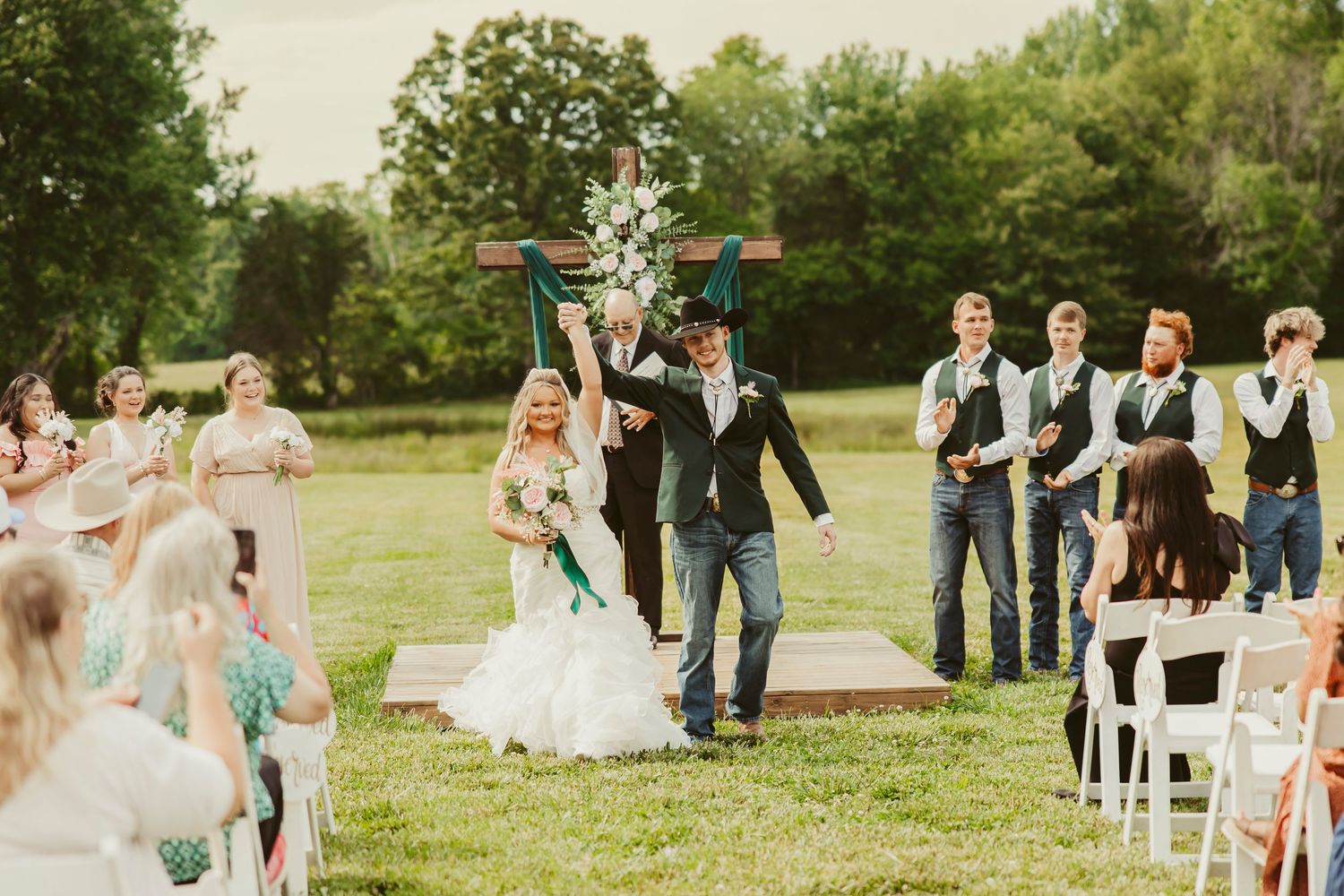 Joyful bride and groom celebrate after outdoor wedding ceremony on green lawn with wedding party and guests cheering.