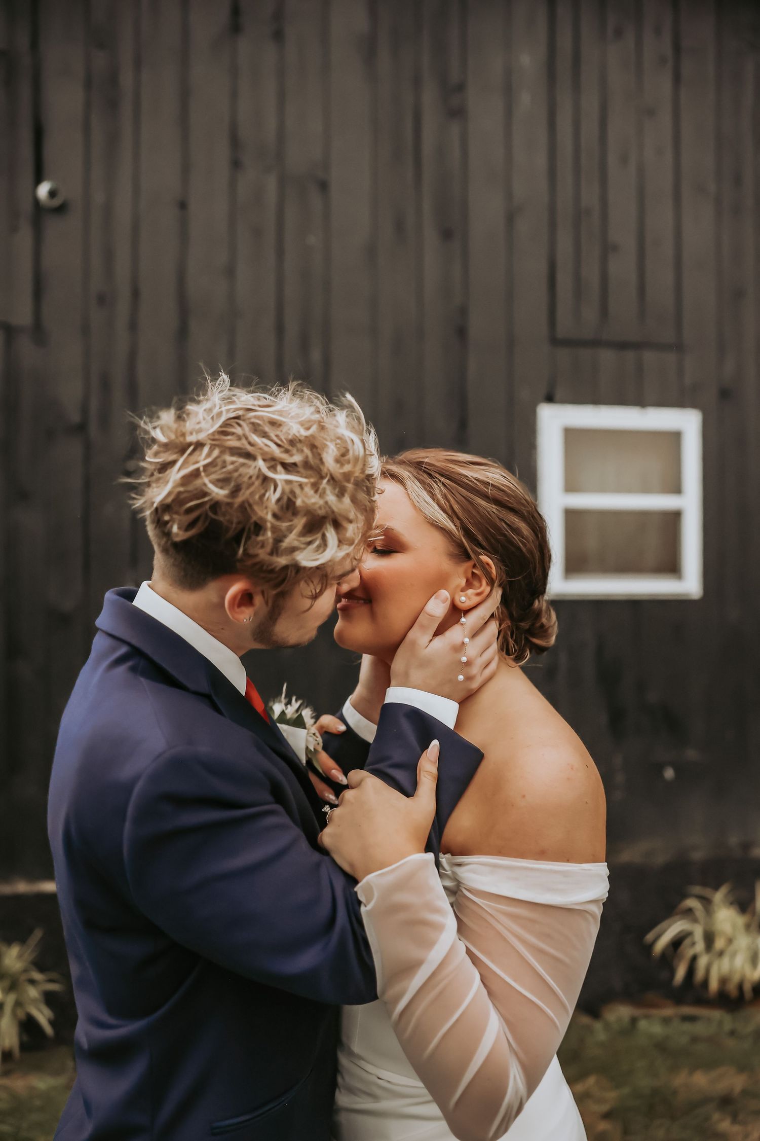An intimate outdoor wedding portrait of a couple sharing a tender moment against a dark barn wall.