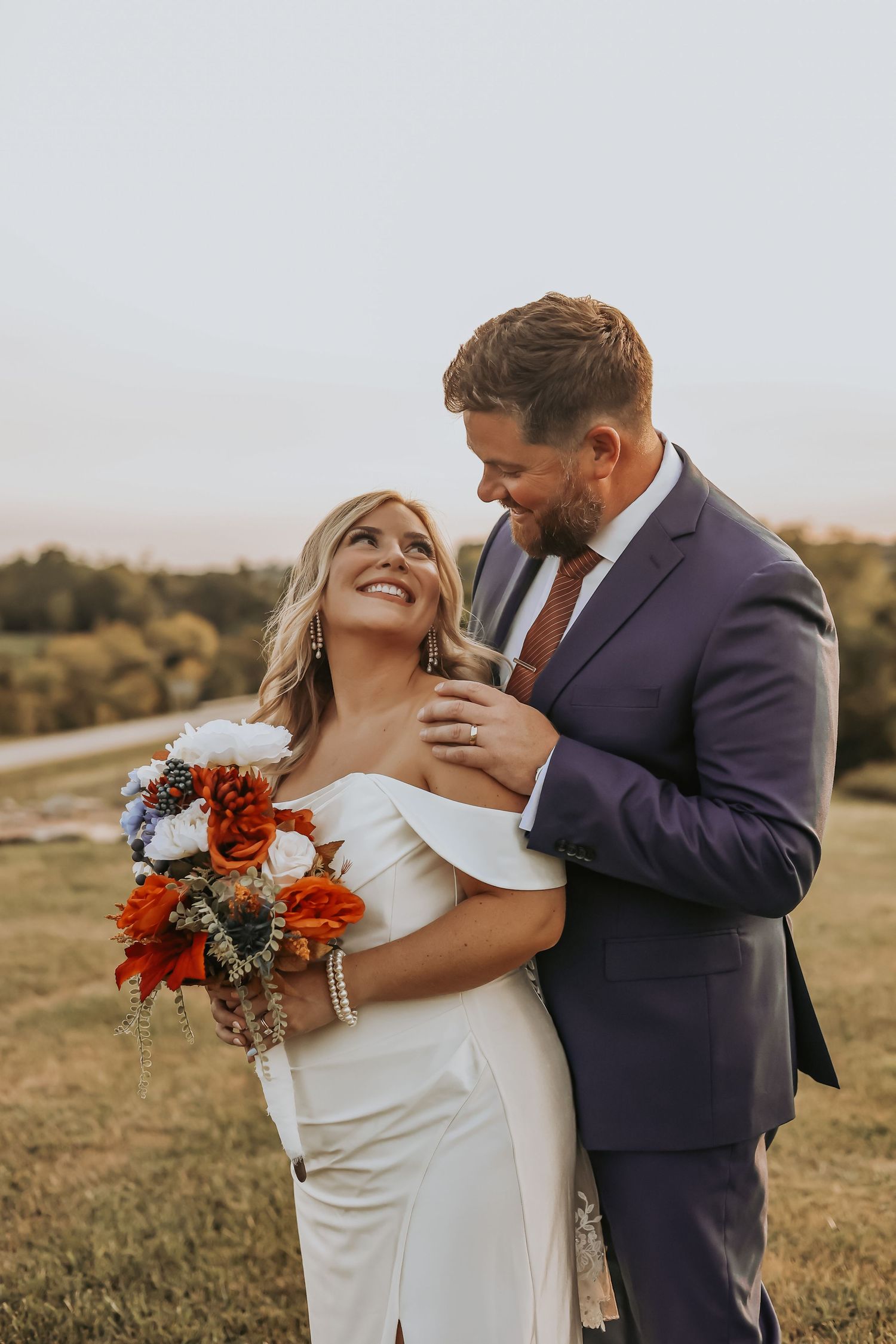 Newlywed couple embraces outdoors with bride holding vibrant red bouquet at autumn wedding.