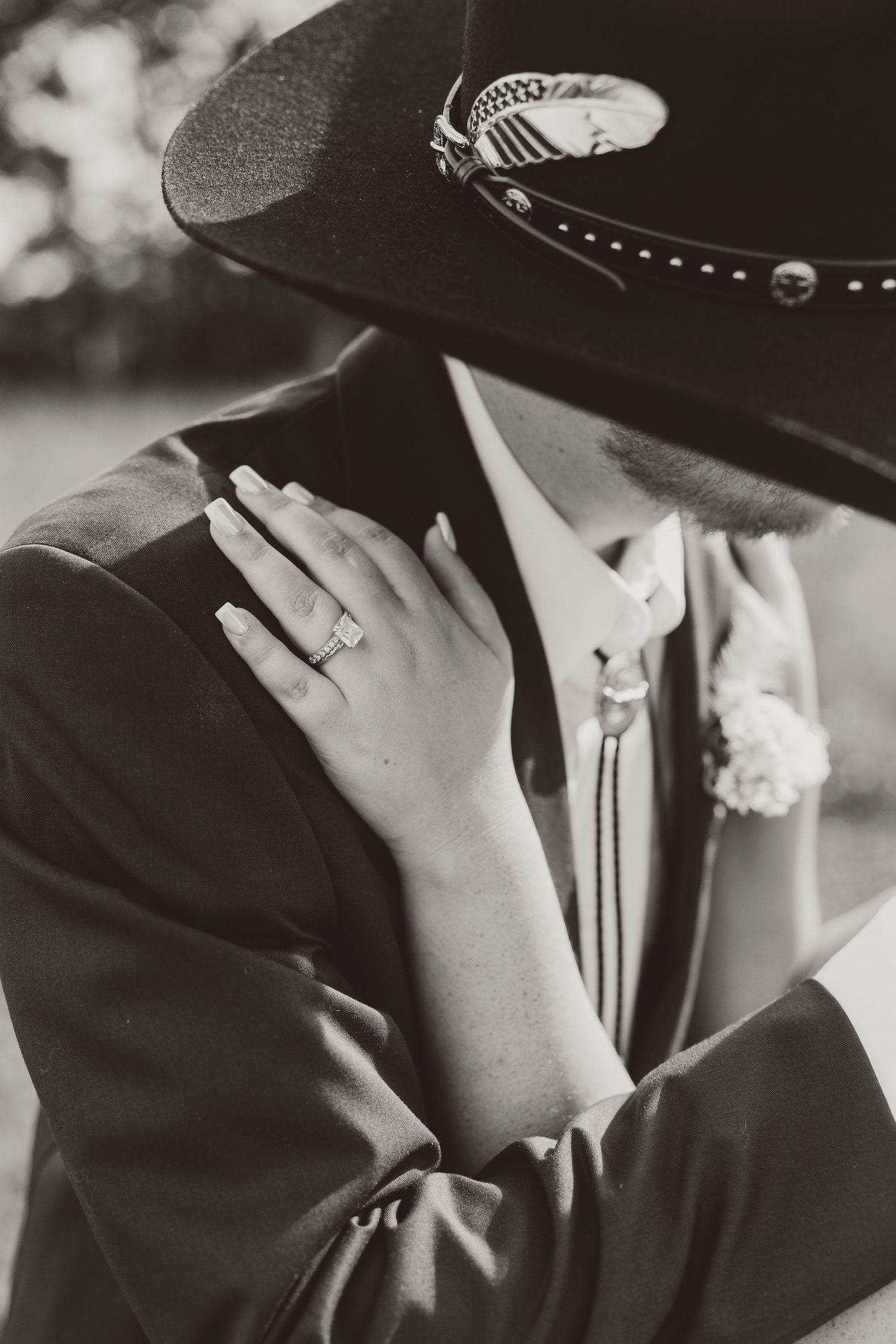Black and white photo of a couple embracing, showcasing an engagement ring, cowboy hat, bolo tie, and boutonniere.
