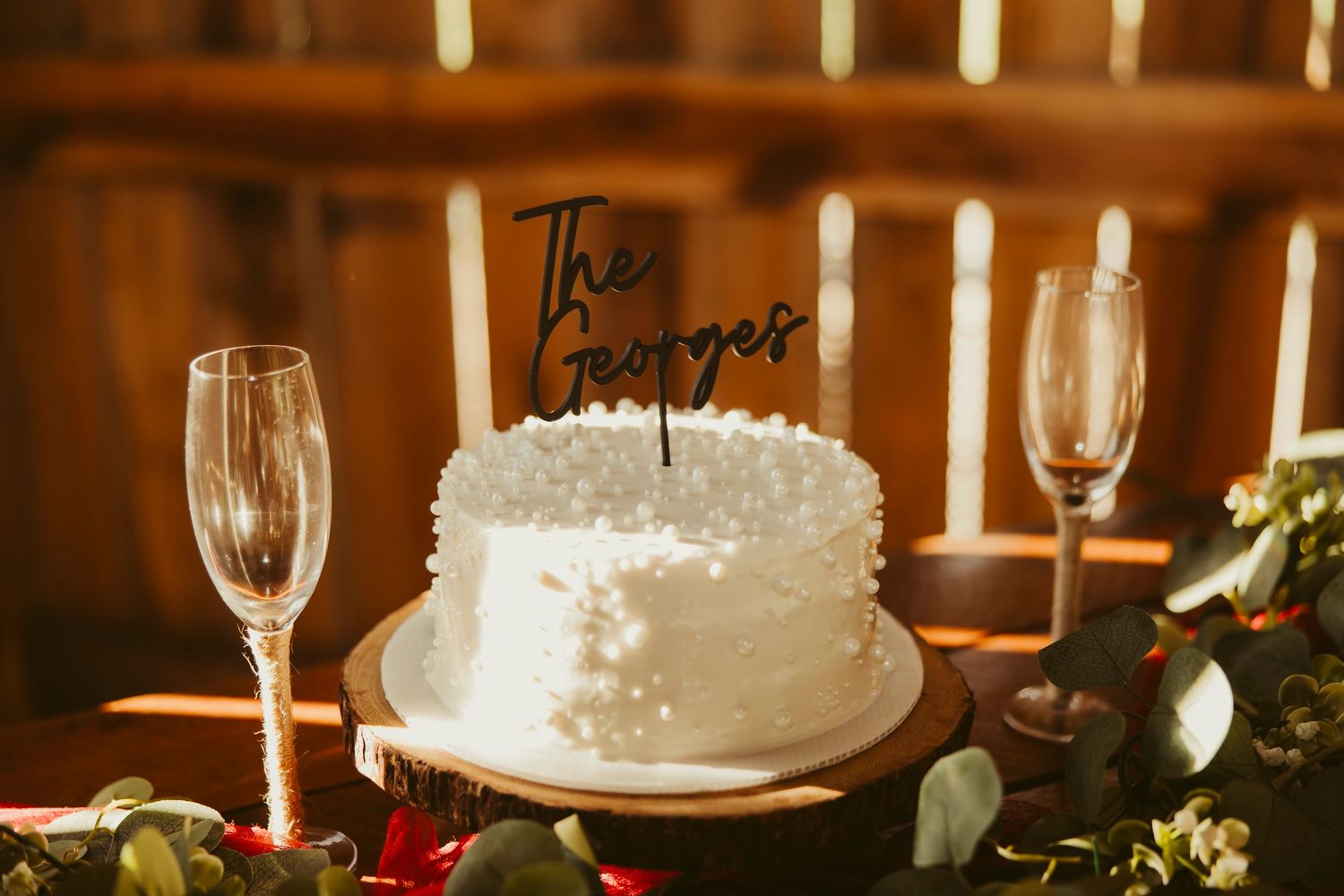 White wedding cake with The Georges topper flanked by champagne flutes and candles on a rustic wooden table.