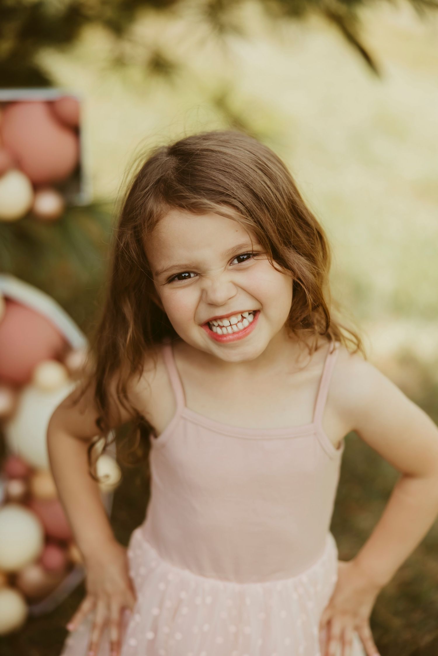 A joyful young girl in a pink dress smiles broadly outdoors near colorful balloons in soft natural light.