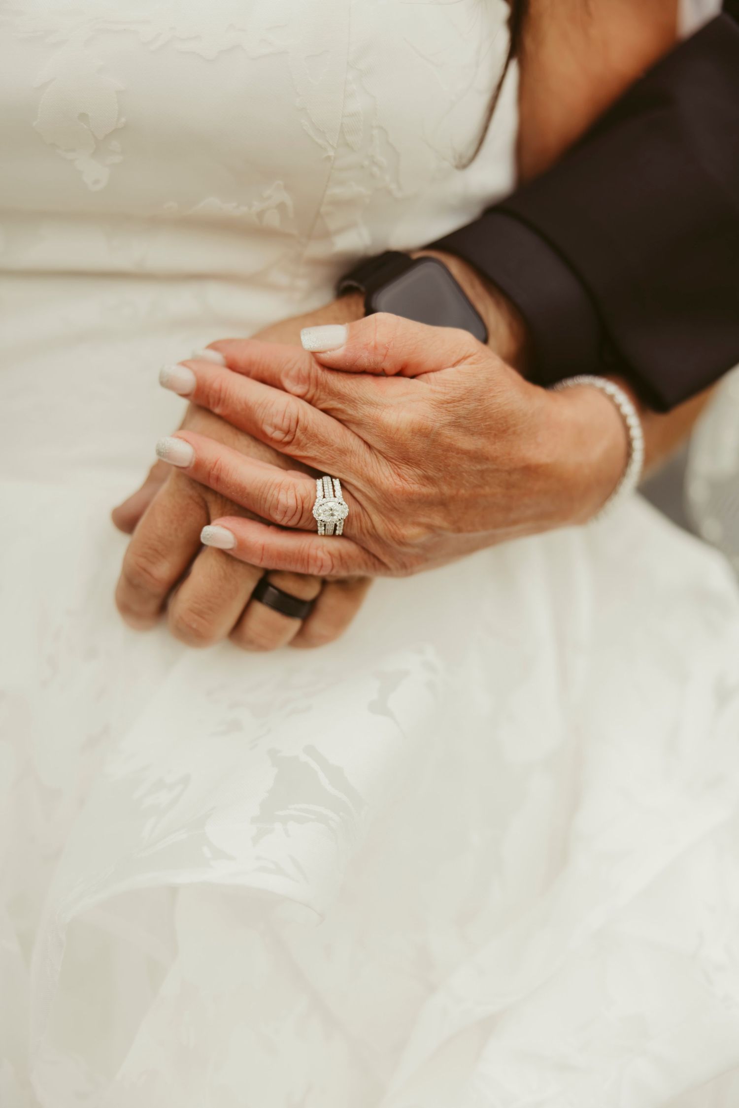 Close-up of bride and groom clasped hands showing diamond wedding ring set and black silicone band on white bridal gown.