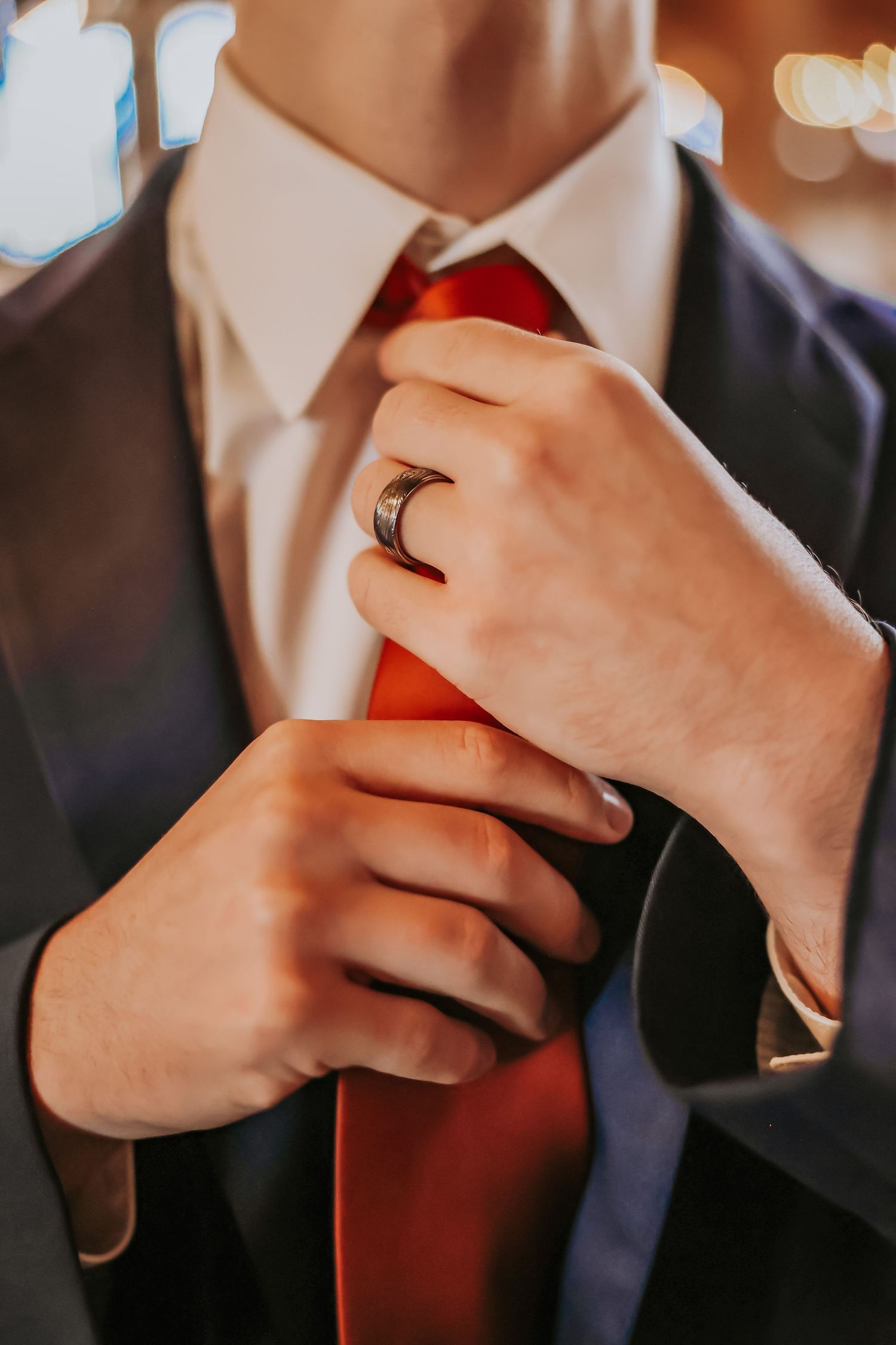 Close-up of groom adjusting bright red tie with wedding band visible.