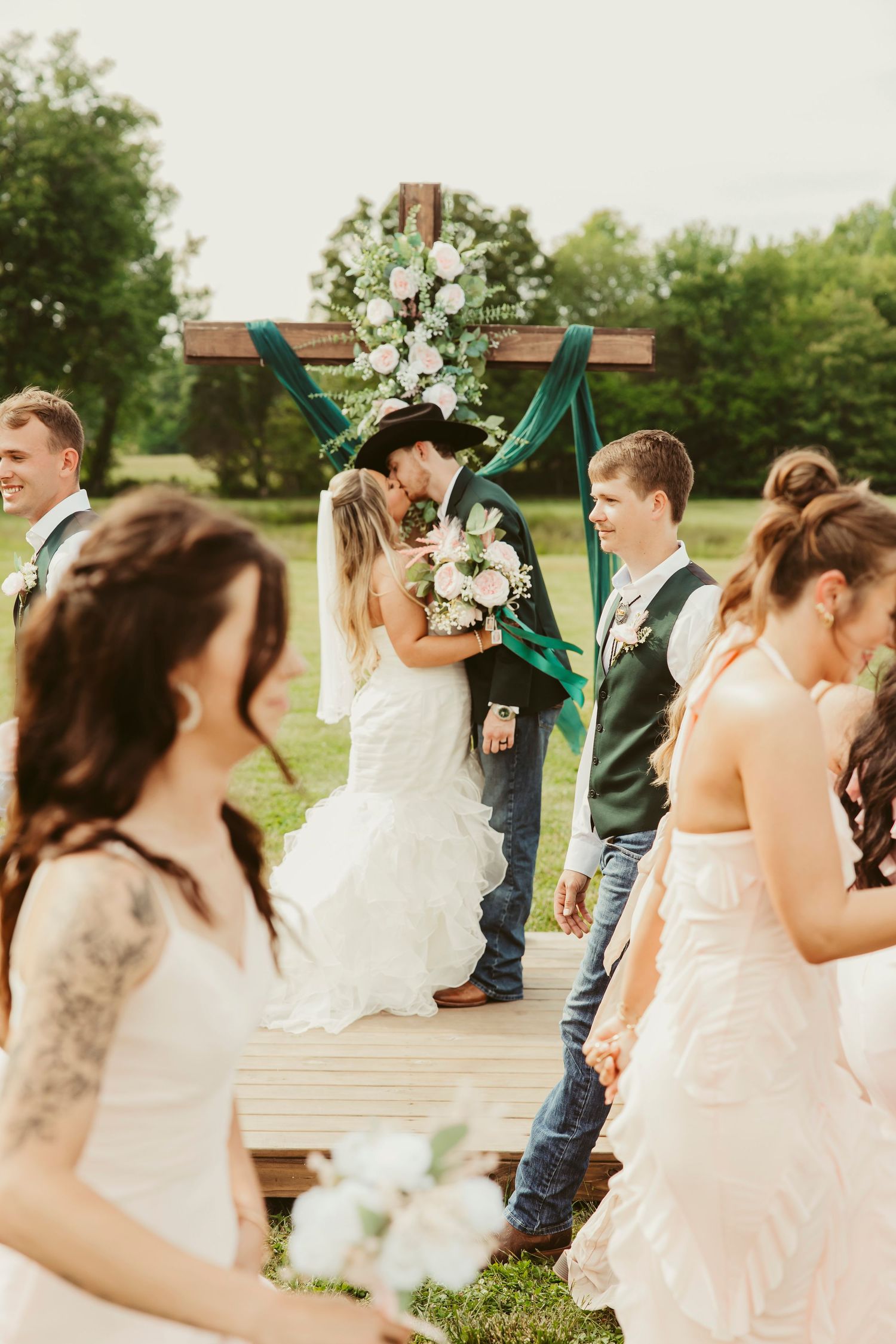 Bride and groom share first kiss at rustic outdoor wedding ceremony with wooden cross arch and green draping.