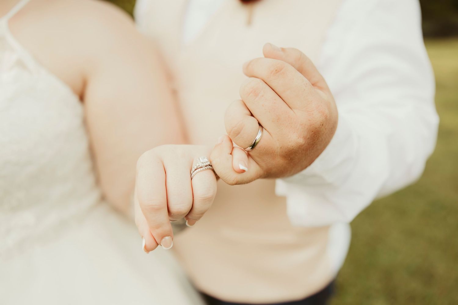 Newlywed couple holding pinkies showing wedding rings, bride in white lace gown and groom in white shirt.