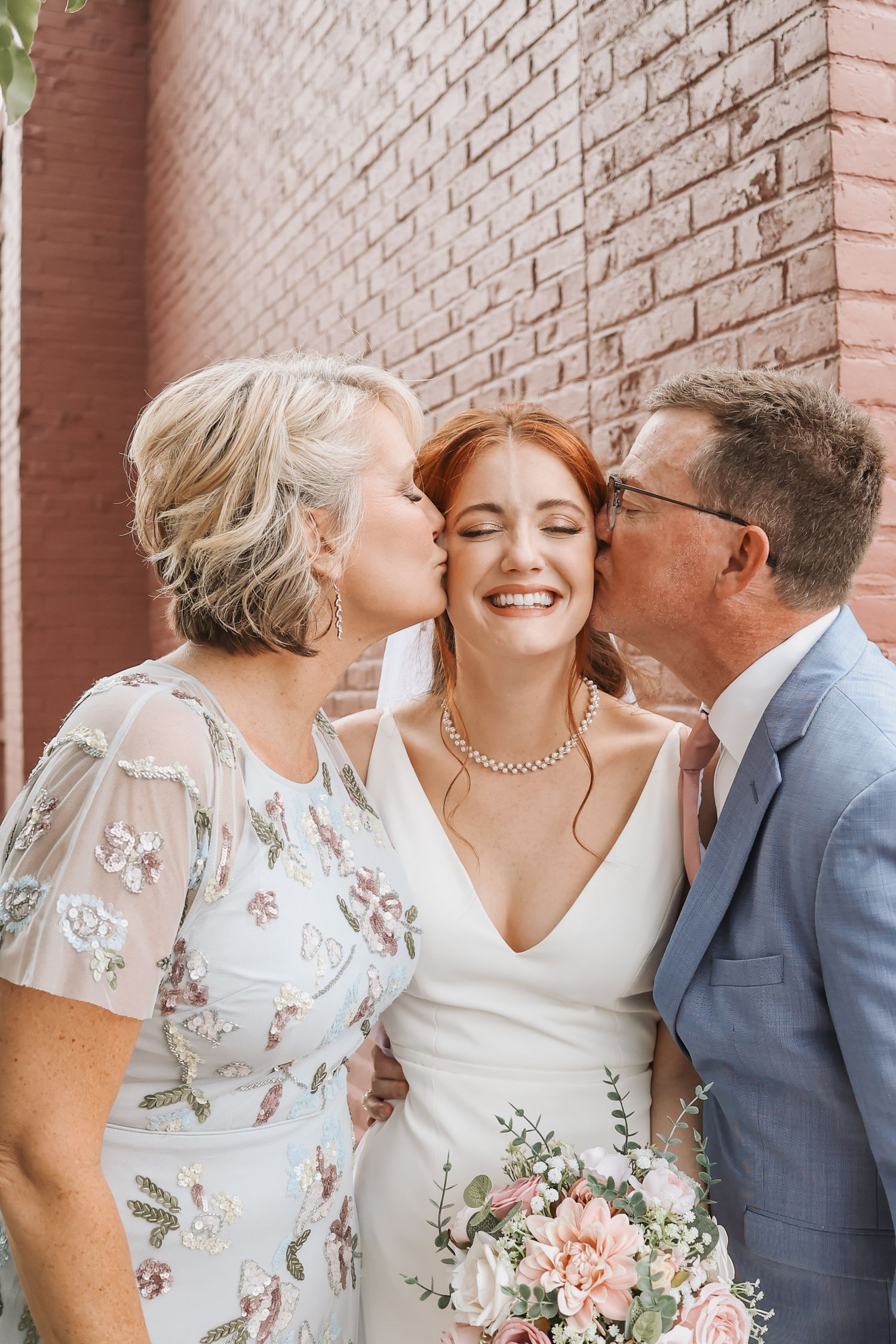 A tender wedding moment captured against a pink brick wall with a bouquet of blush roses.