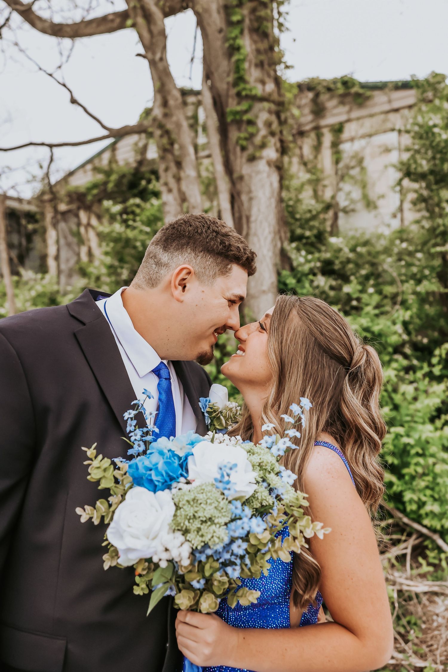 A couple shares a romantic moment while holding a beautiful blue and white wedding bouquet outdoors.