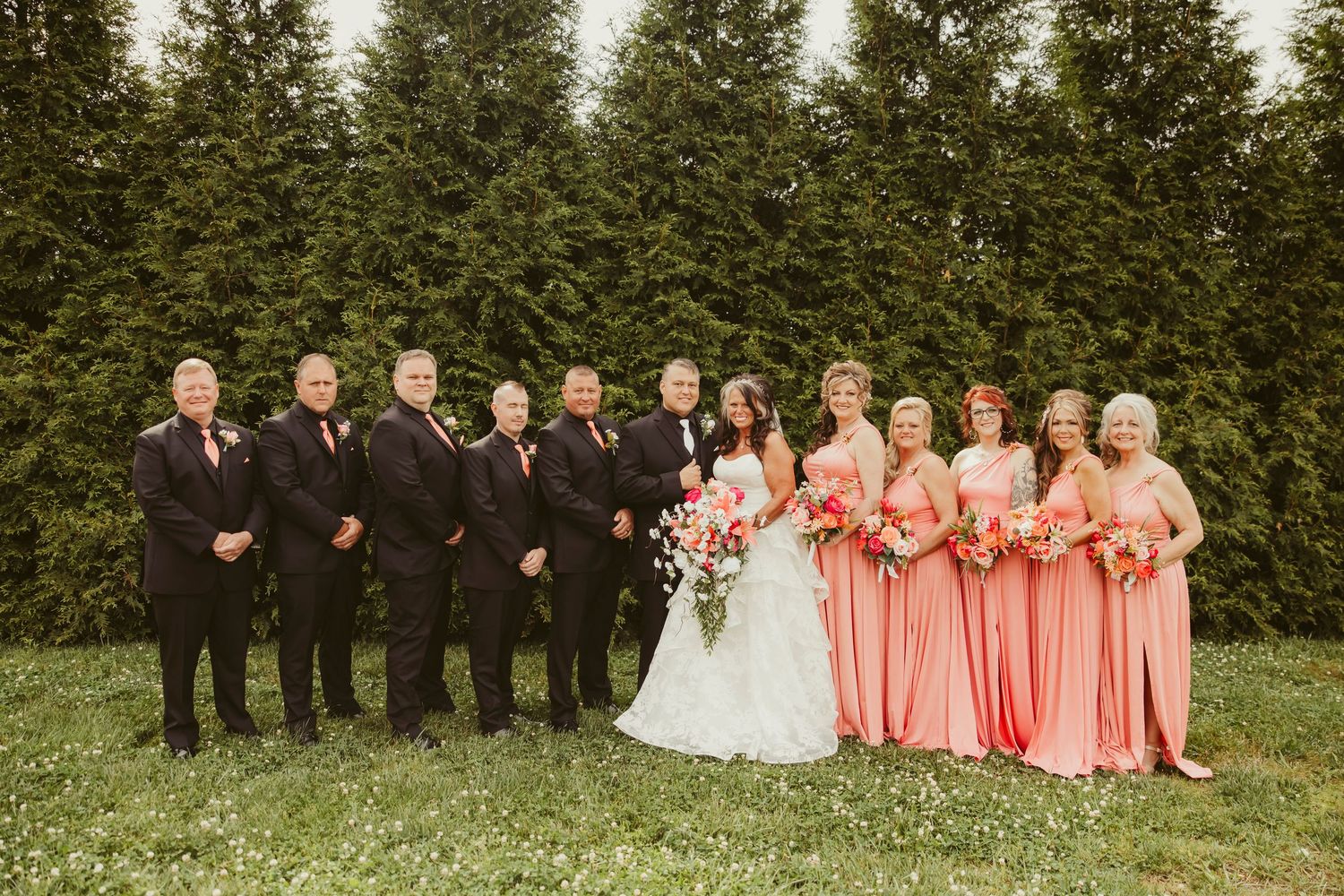 Wedding party poses outdoors, groomsmen in black suits and bridesmaids in coral gowns holding bouquets, with evergreen trees behind.