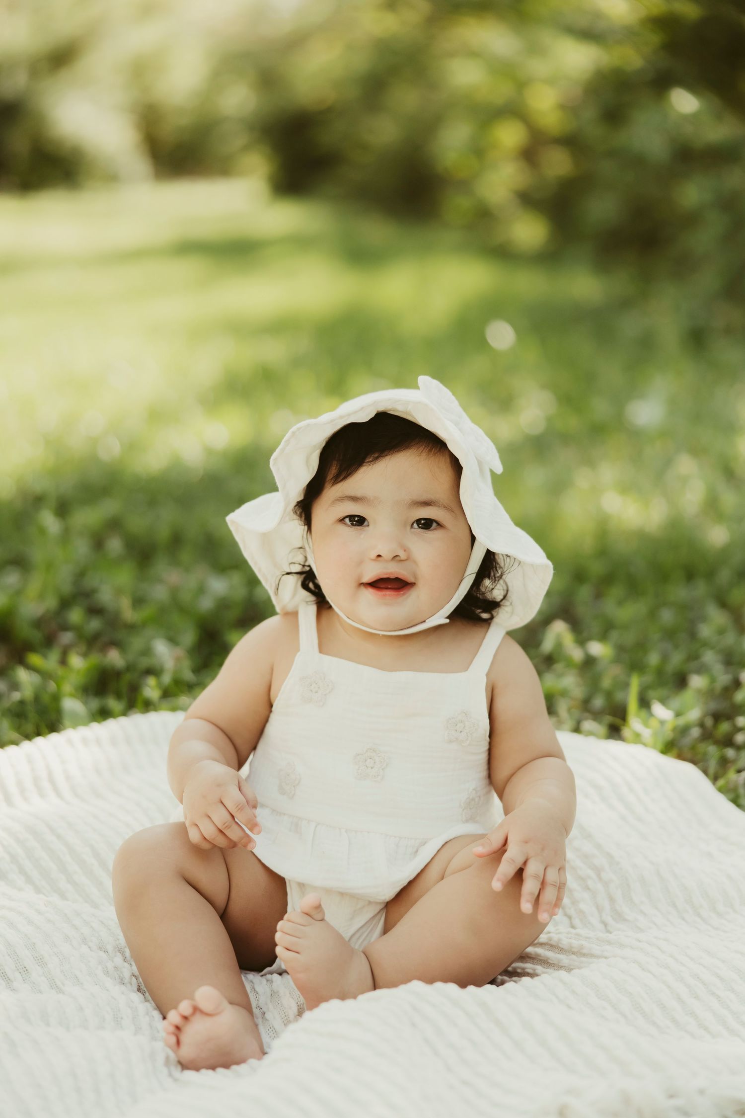 Adorable baby girl in white romper and bonnet hat sitting on blanket outdoors in green garden.