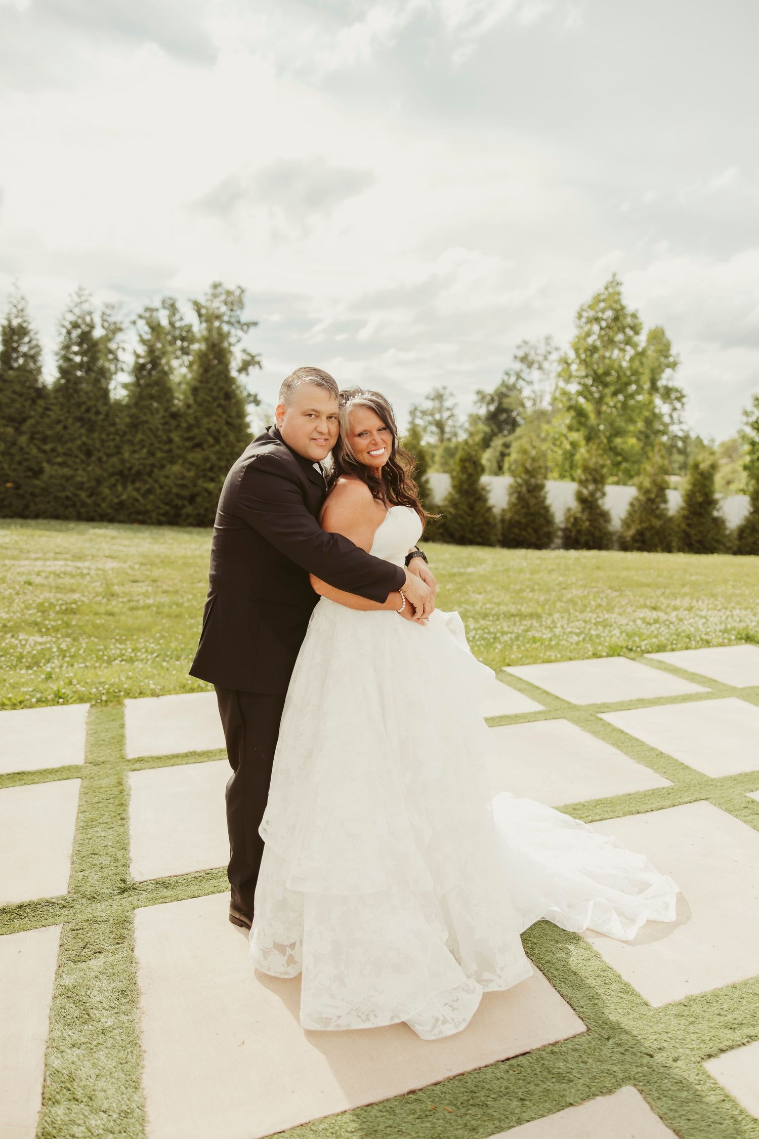 Bride and groom embrace outdoors on a checkered stone patio, surrounded by lush greenery on their wedding day.