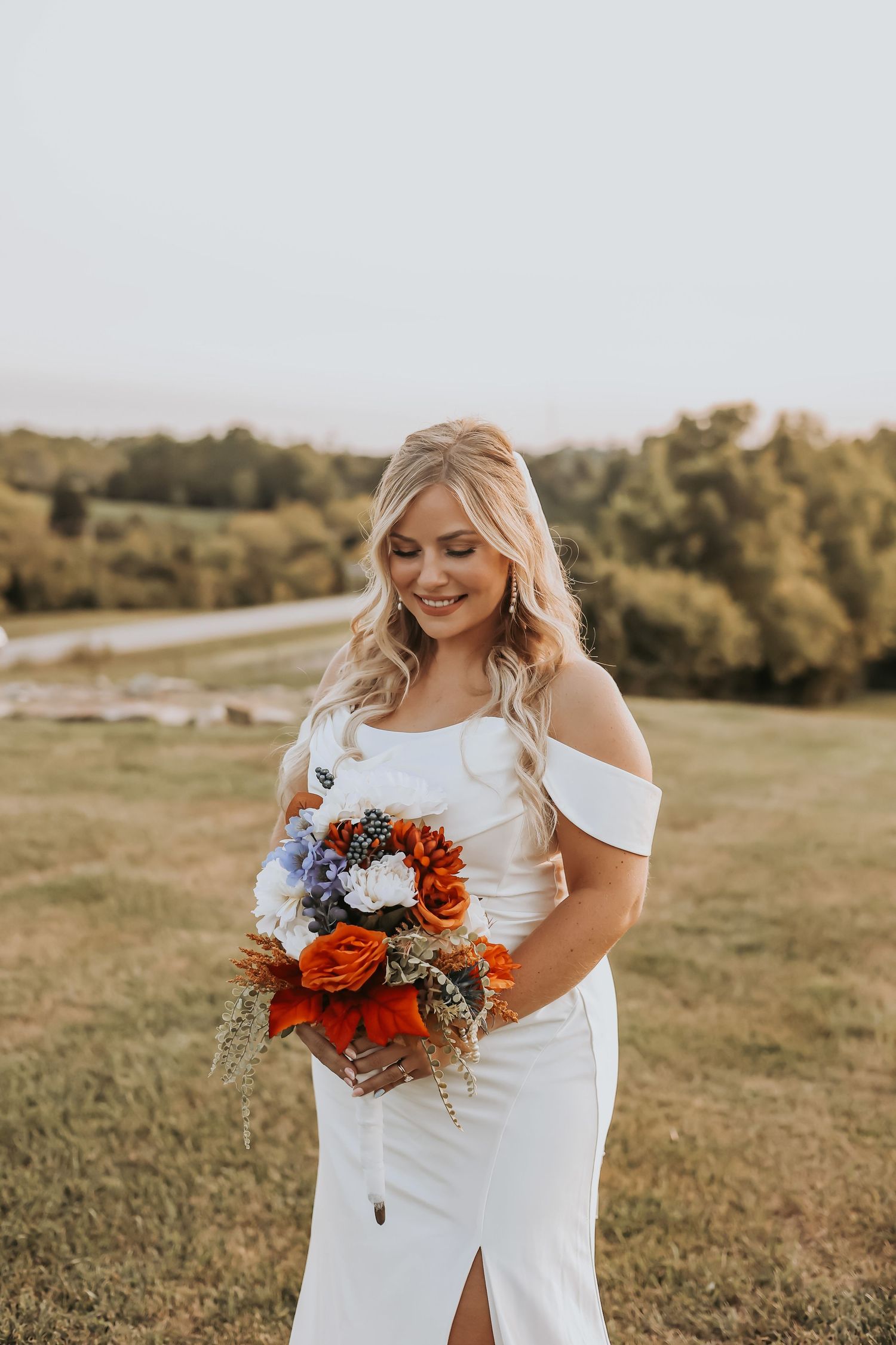 Bride in off-shoulder white dress holds orange and blue bouquet in outdoor setting.