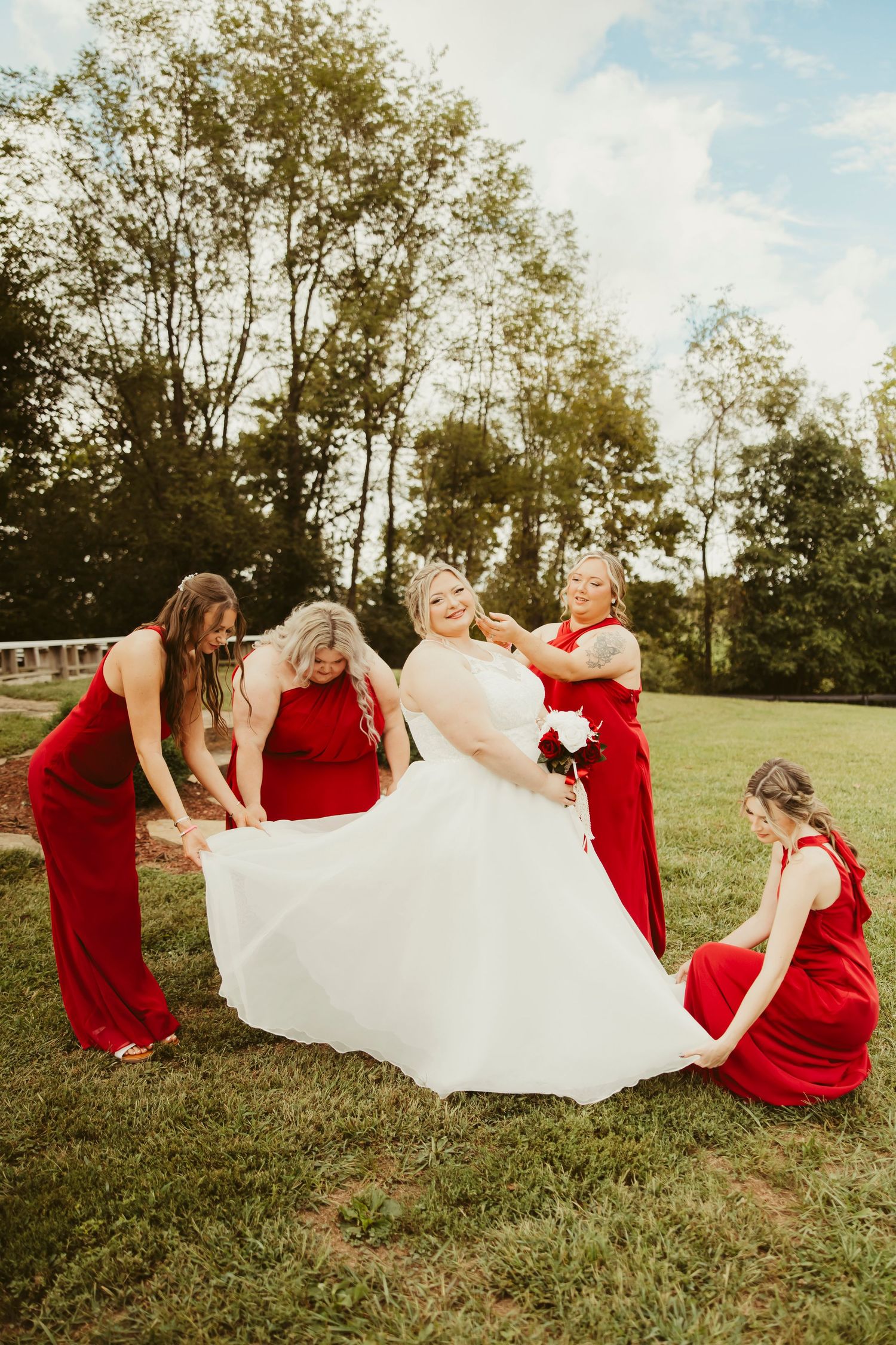 Bride in white gown twirling with four bridesmaids in red dresses on green lawn with trees in background.