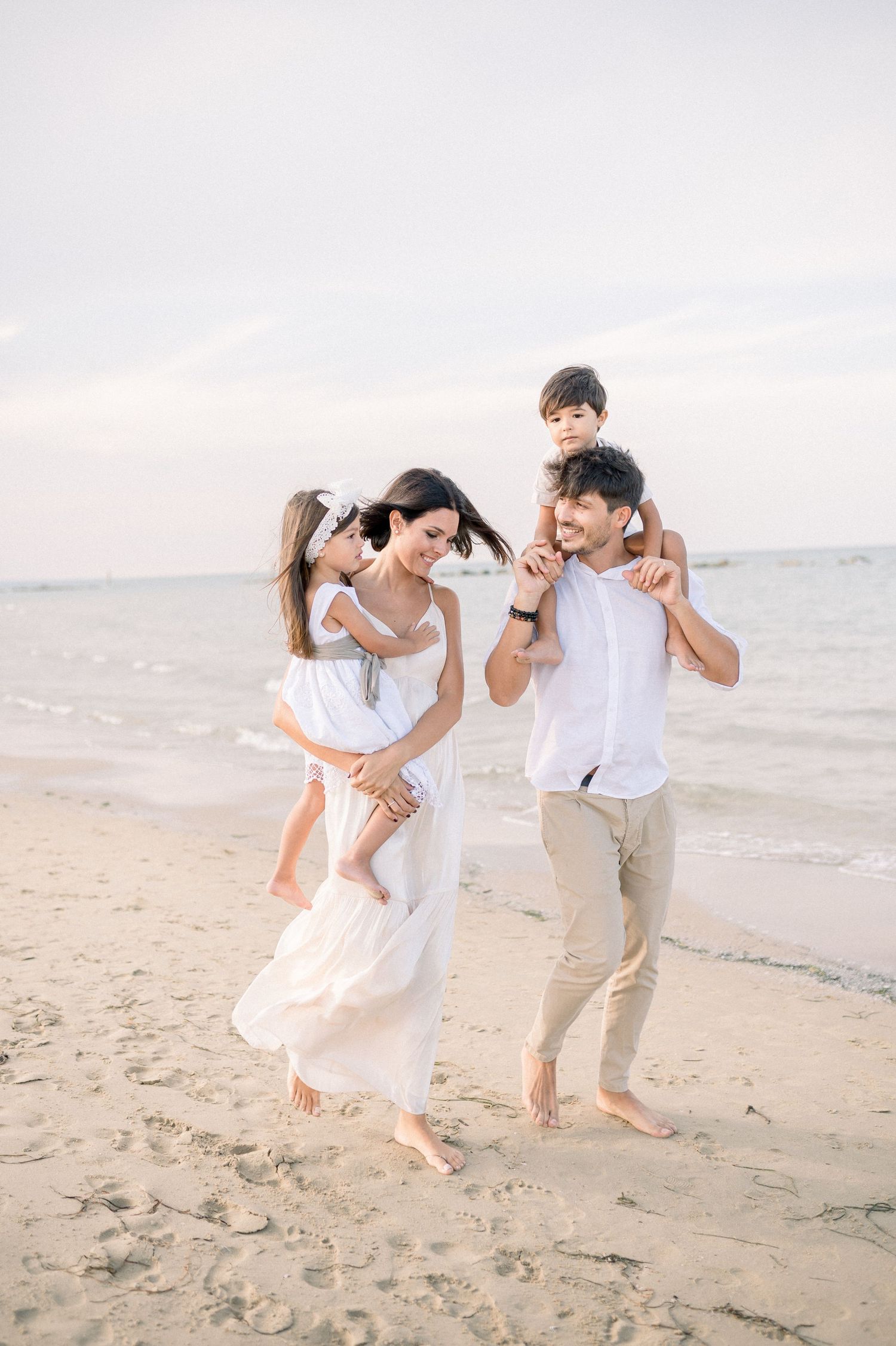 Family playing and jumping together on beach wearing white casual outfits.