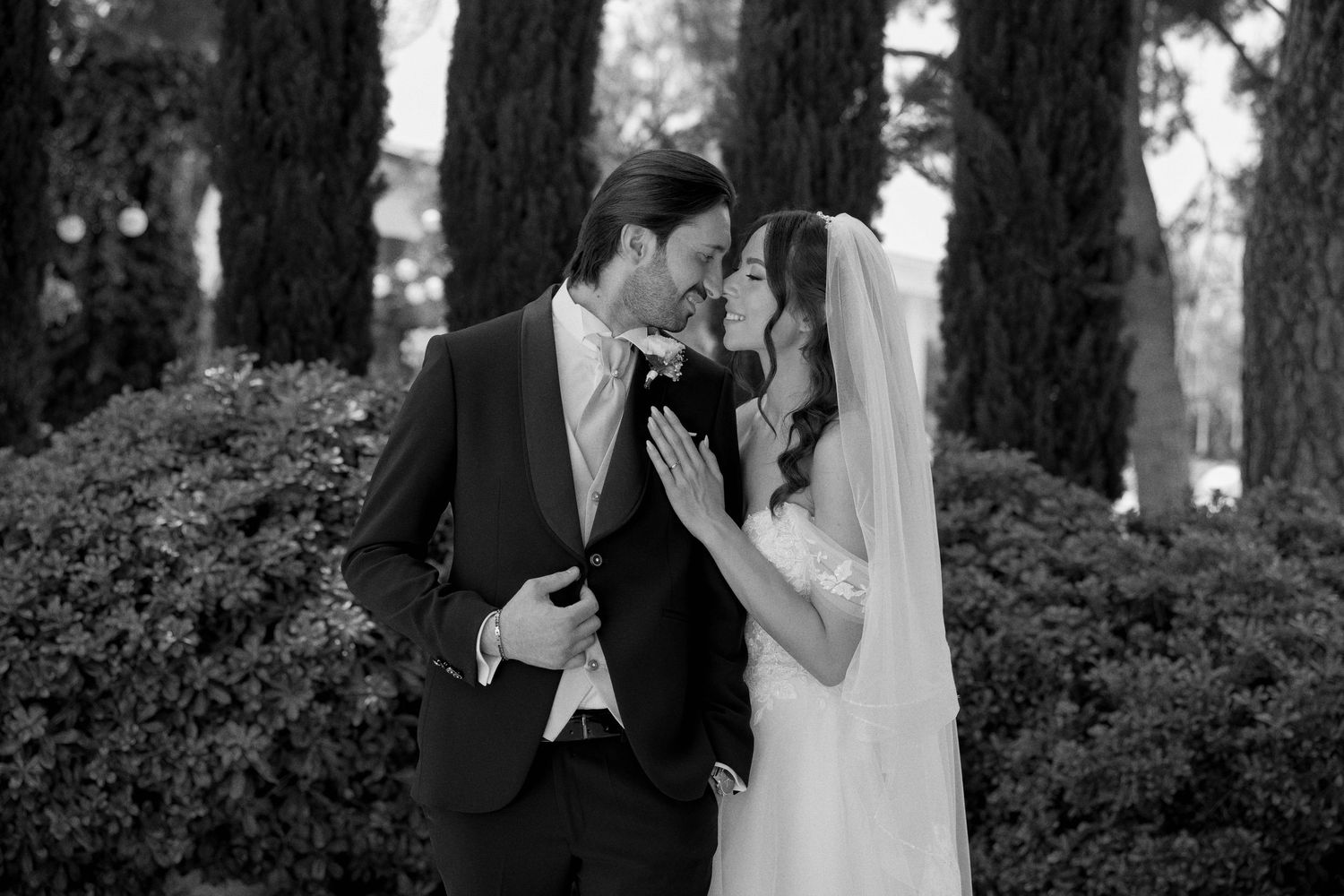 Newlyweds embrace in a lush garden setting with evergreen trees in the background.