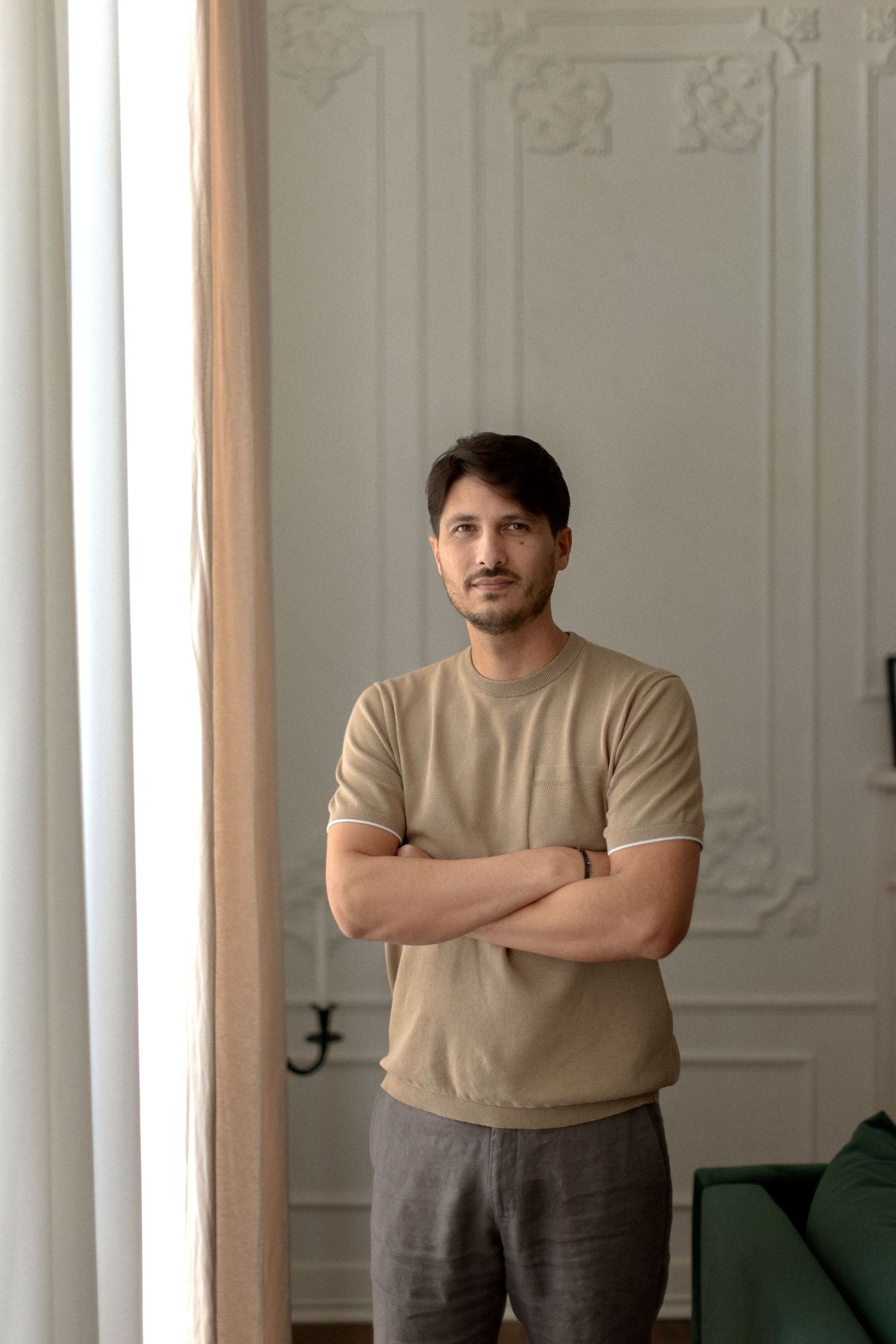 Person in beige t-shirt stands with arms crossed in front of white ornate wall in bright interior setting.