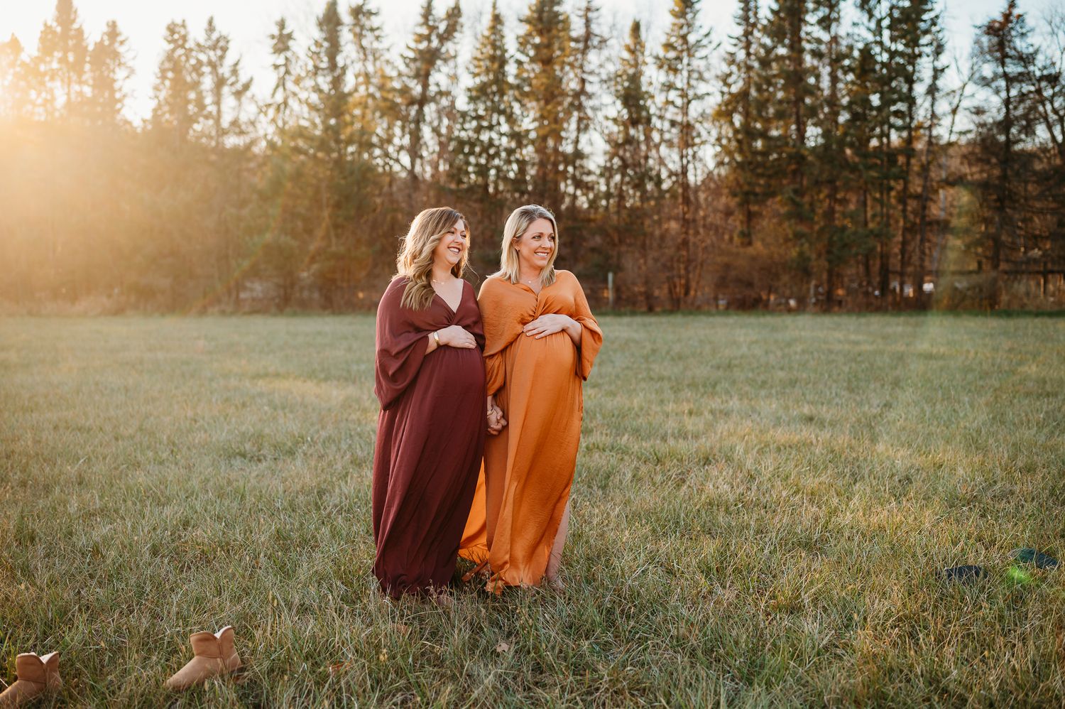 Two people in flowing autumn-colored dresses walk together in a sunset-lit meadow with evergreen trees in the background.