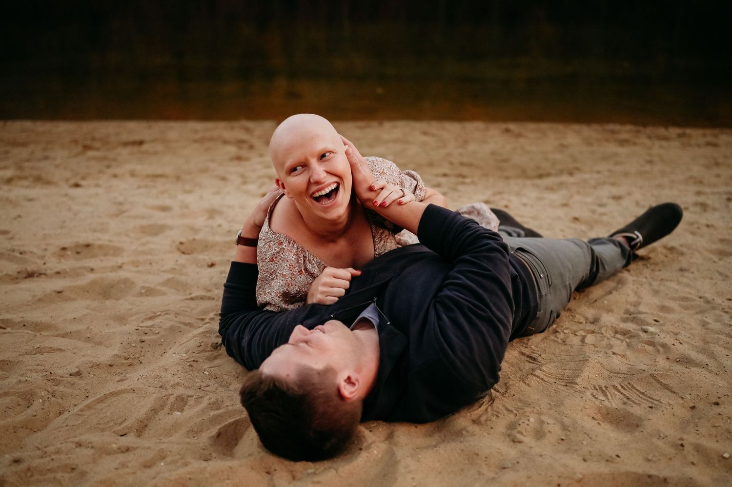 A romantic couple embraces while lying on sandy beach during sunset photoshoot.