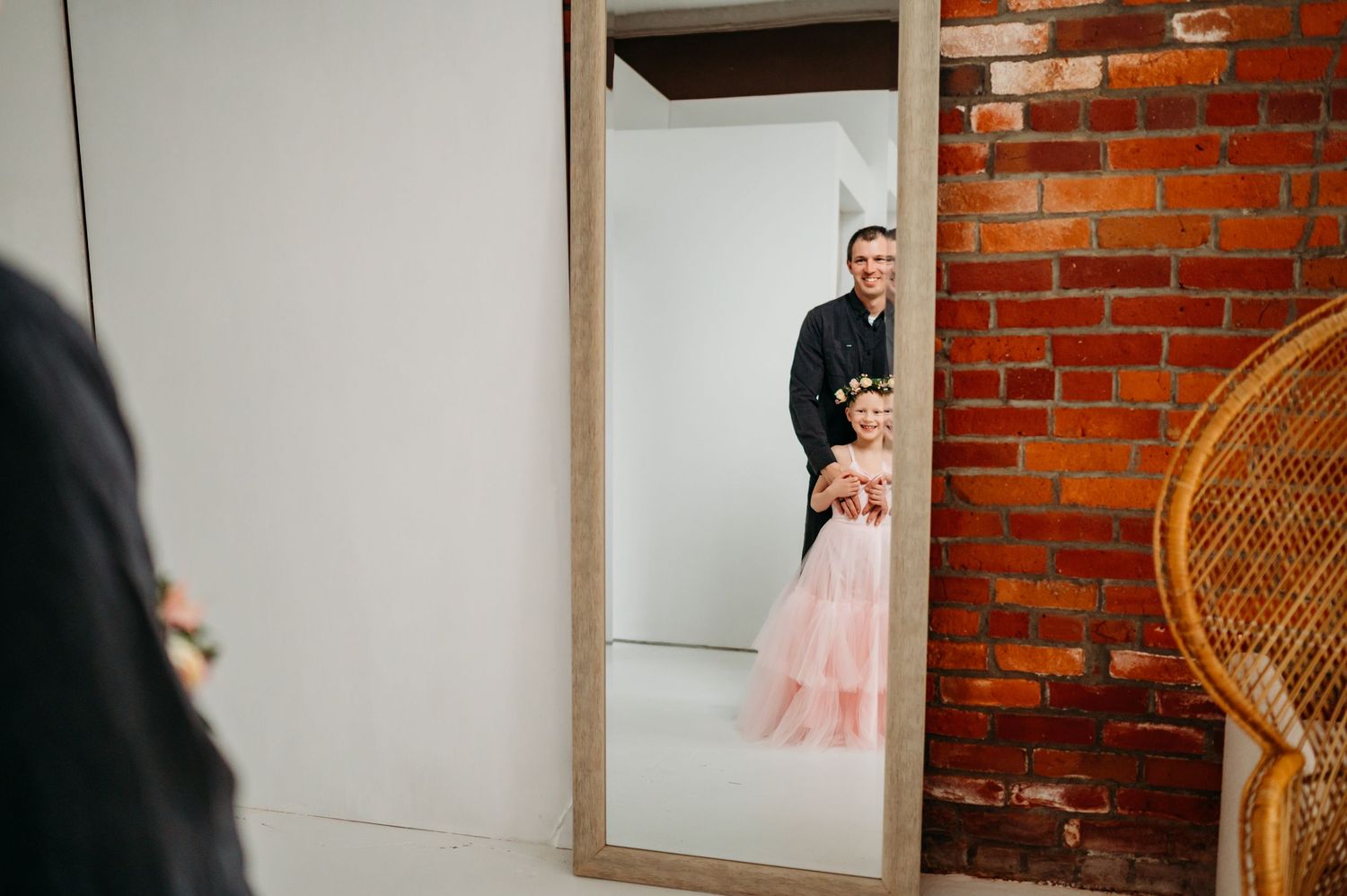 A reflection in a mirror shows two figures in a room with exposed brick walls and a rattan chair.