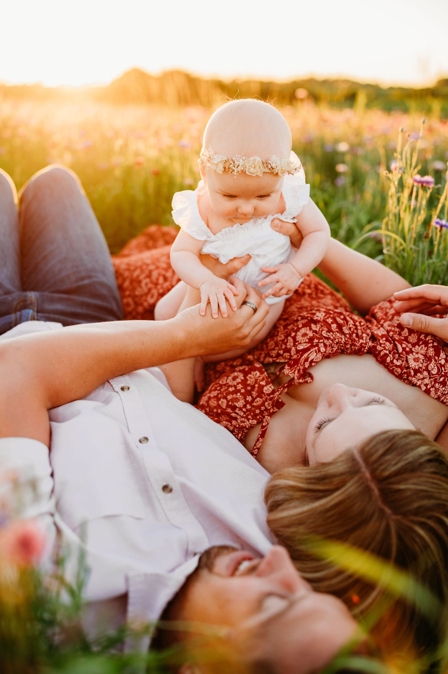 Parents lying in wildflower meadow at sunset holding baby wearing floral crown during golden hour photo session.