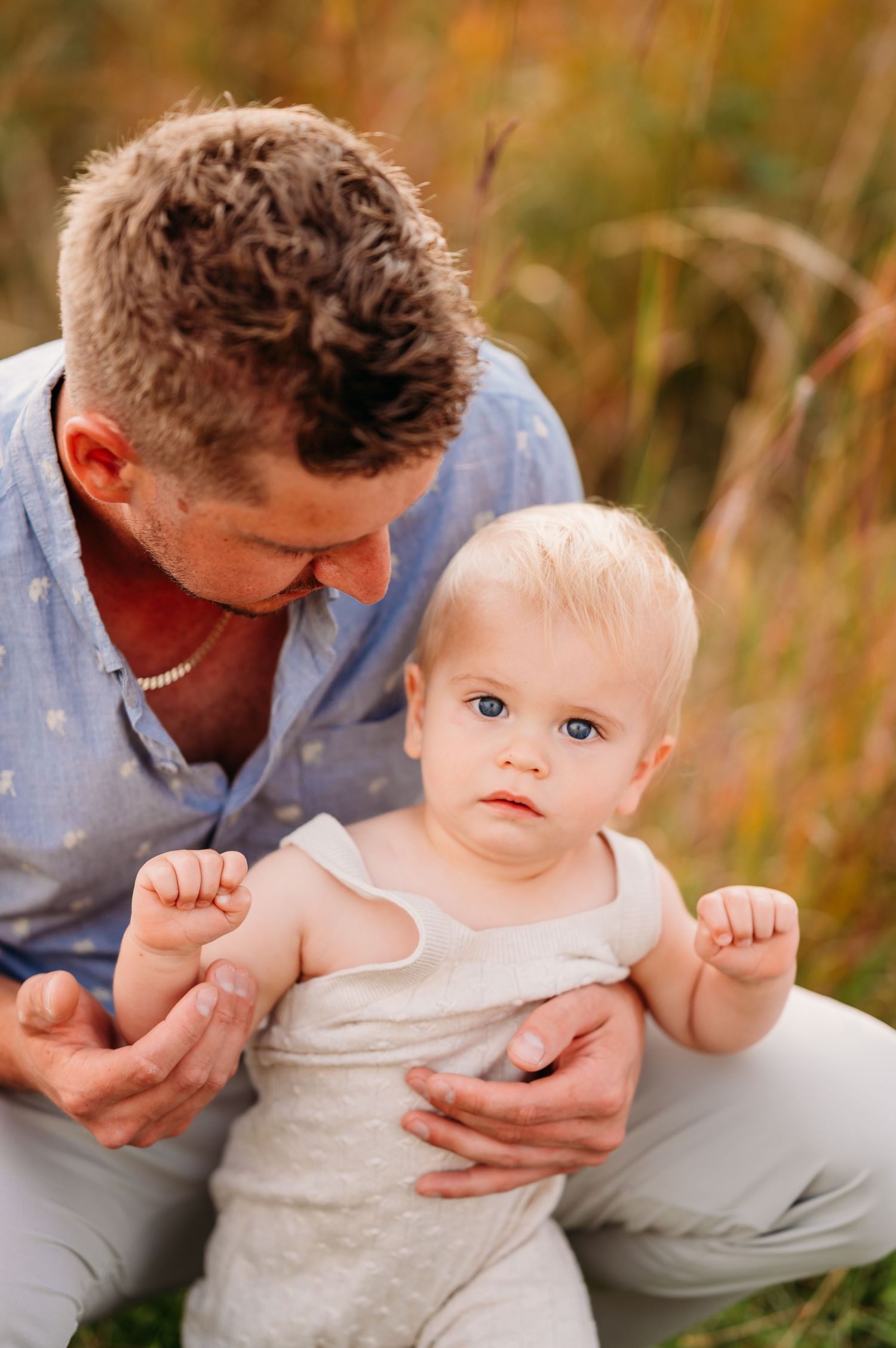 A tender moment outdoors as a baby in white clothing is held in a grassy field with golden autumn colors.