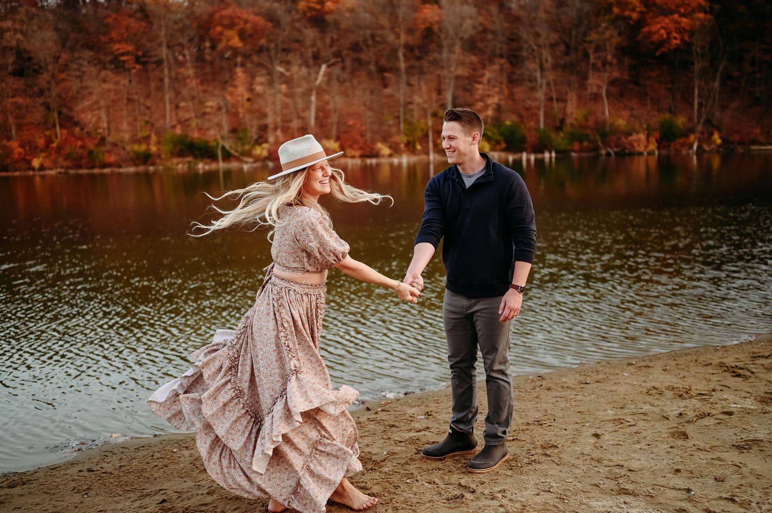 A couple dances playfully on a beach with autumn foliage reflecting in the lake behind them.
