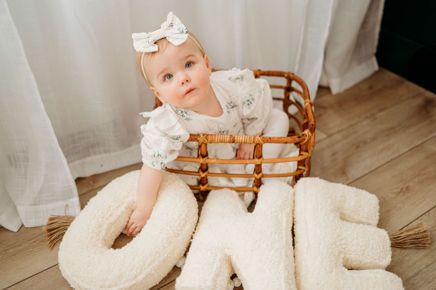Baby in white dress sits in woven wooden chair with plush ONE letters arranged for first birthday photo.