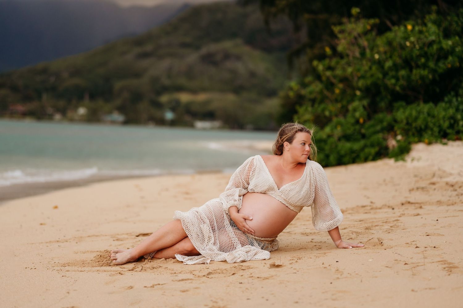A pregnant figure in a flowing white dress reclines on a scenic tropical beach at sunset with mountains in the background.