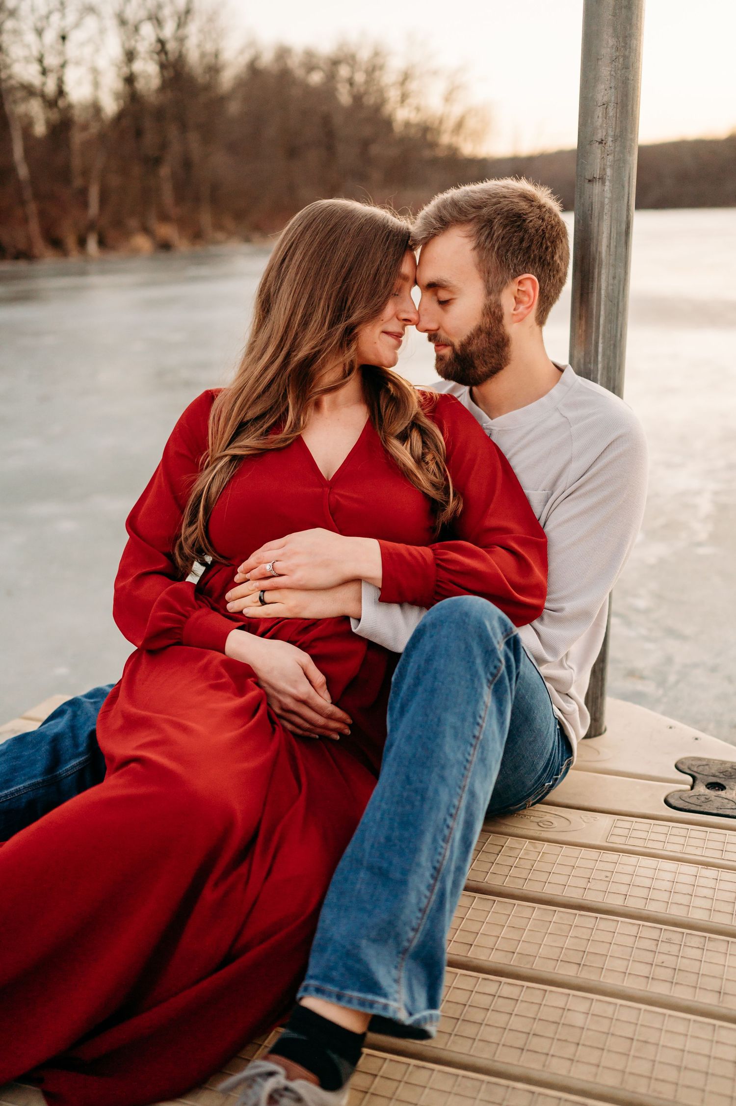 A couple shares an intimate moment on a wooden dock at sunset, with her wearing a flowing red dress.