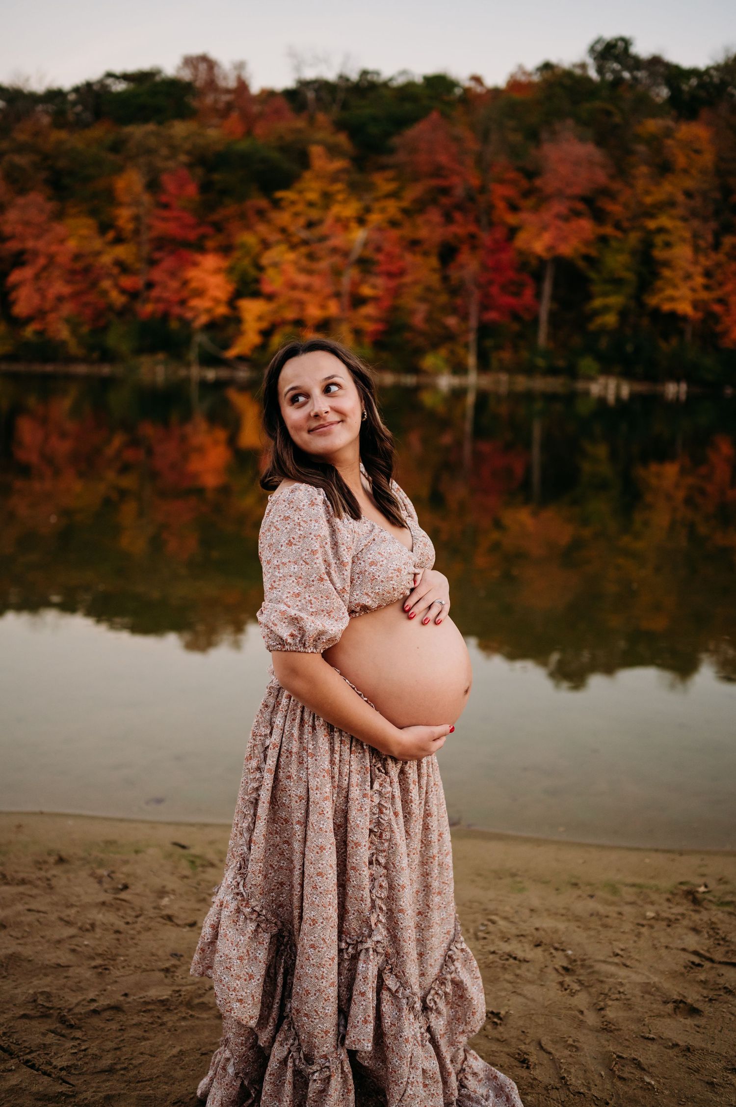 A maternity photo by a lake with fall foliage backdrop showing a woman in a flowy mauve dress holding her baby bump.