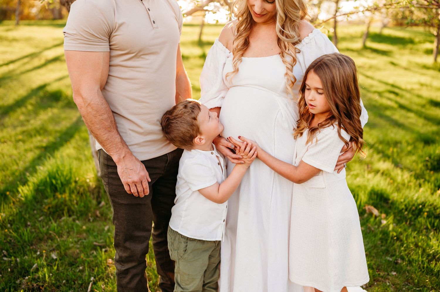 A family stands together in a sunlit meadow wearing white and beige clothing during golden hour.