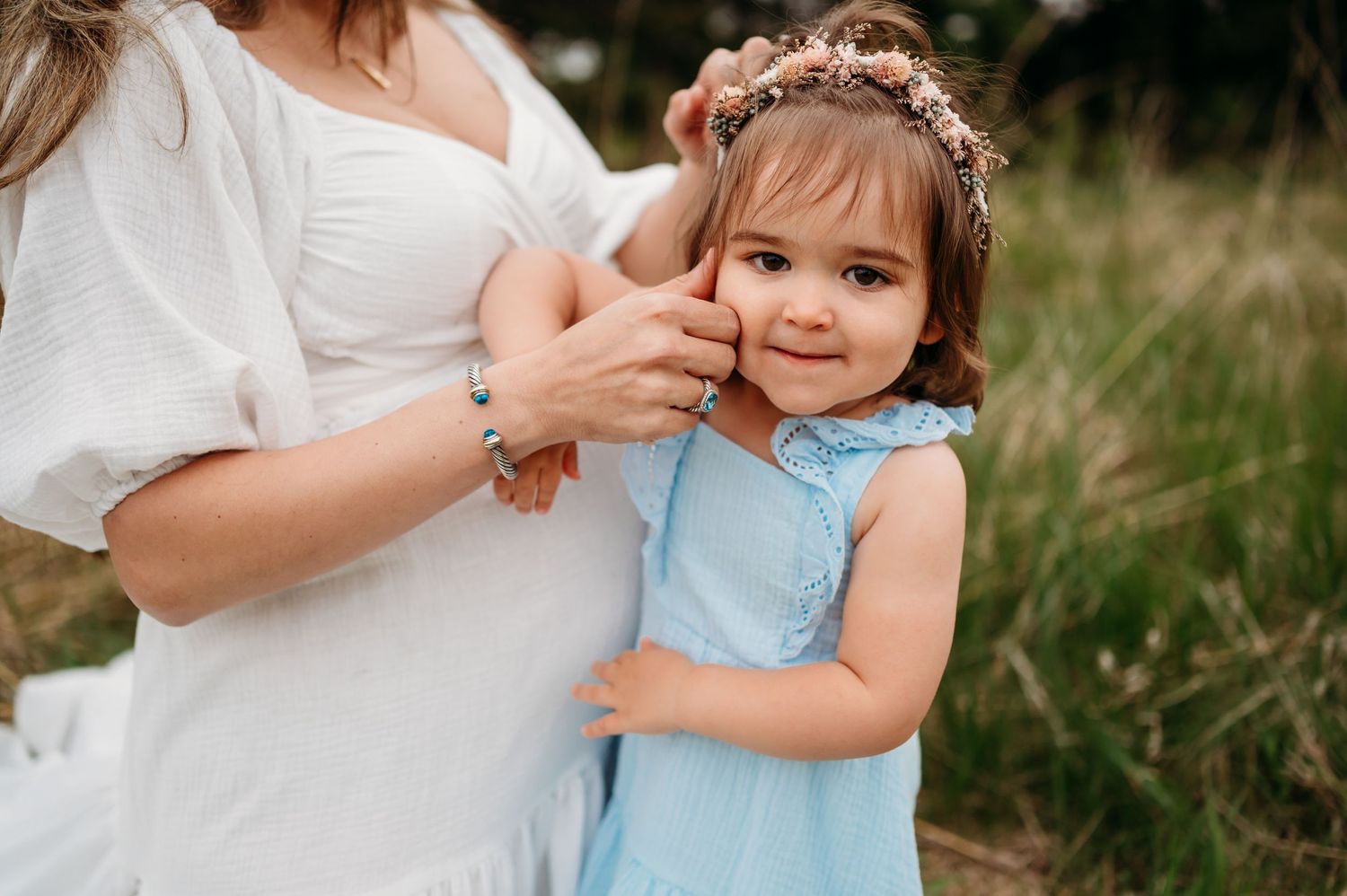 Someone wearing a white dress gently styles small child's hair outdoors in a grassy field setting.