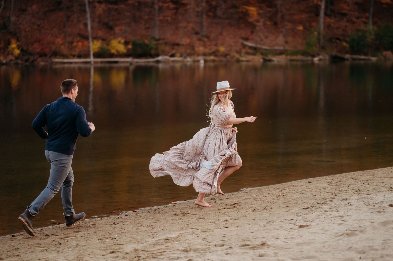 Couple playfully runs and dances along a scenic autumn lakeside path during a romantic photoshoot.