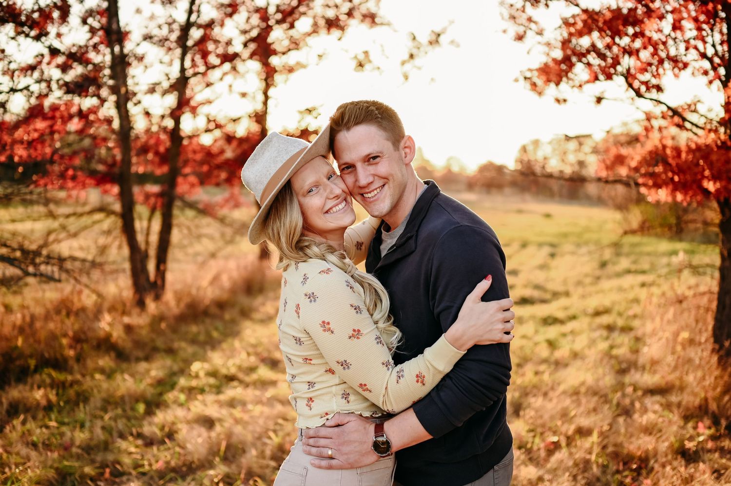 A couple embraces in a romantic autumn setting with golden sunlight and red fall foliage in the background.